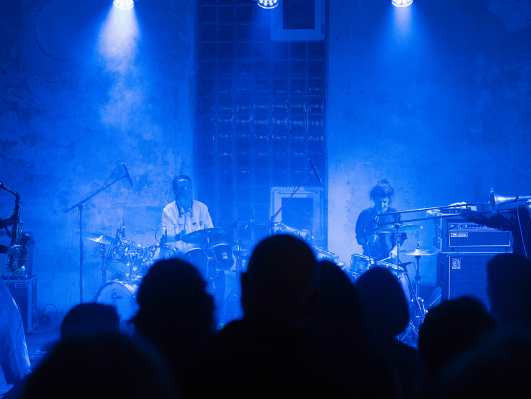 Konsertscene i blått lys, to musikere ved trommer og utstyr, silhuetter av publikum i forgrunnen.