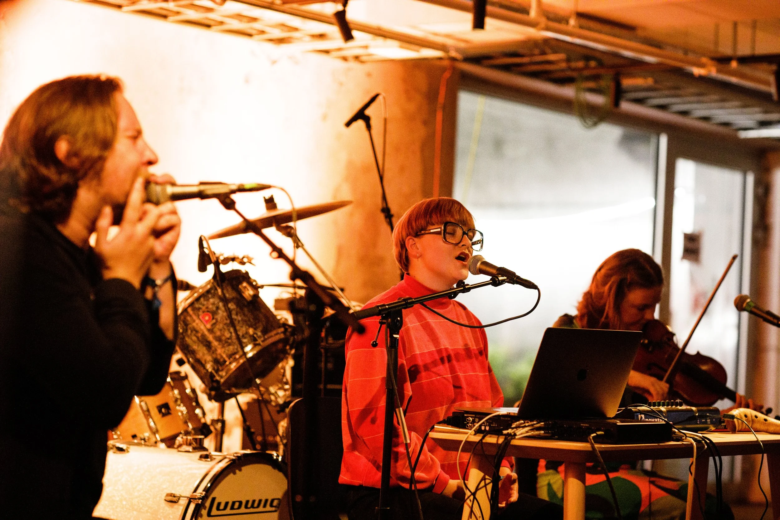 Tre musikere på scene i varmt lys. Til venstre synger en mann i mikrofon, i midten synger en kvinne bak laptop og synth-utstyr, til høyre spiller en fiolinist. Trommesett i bakgrunnen.