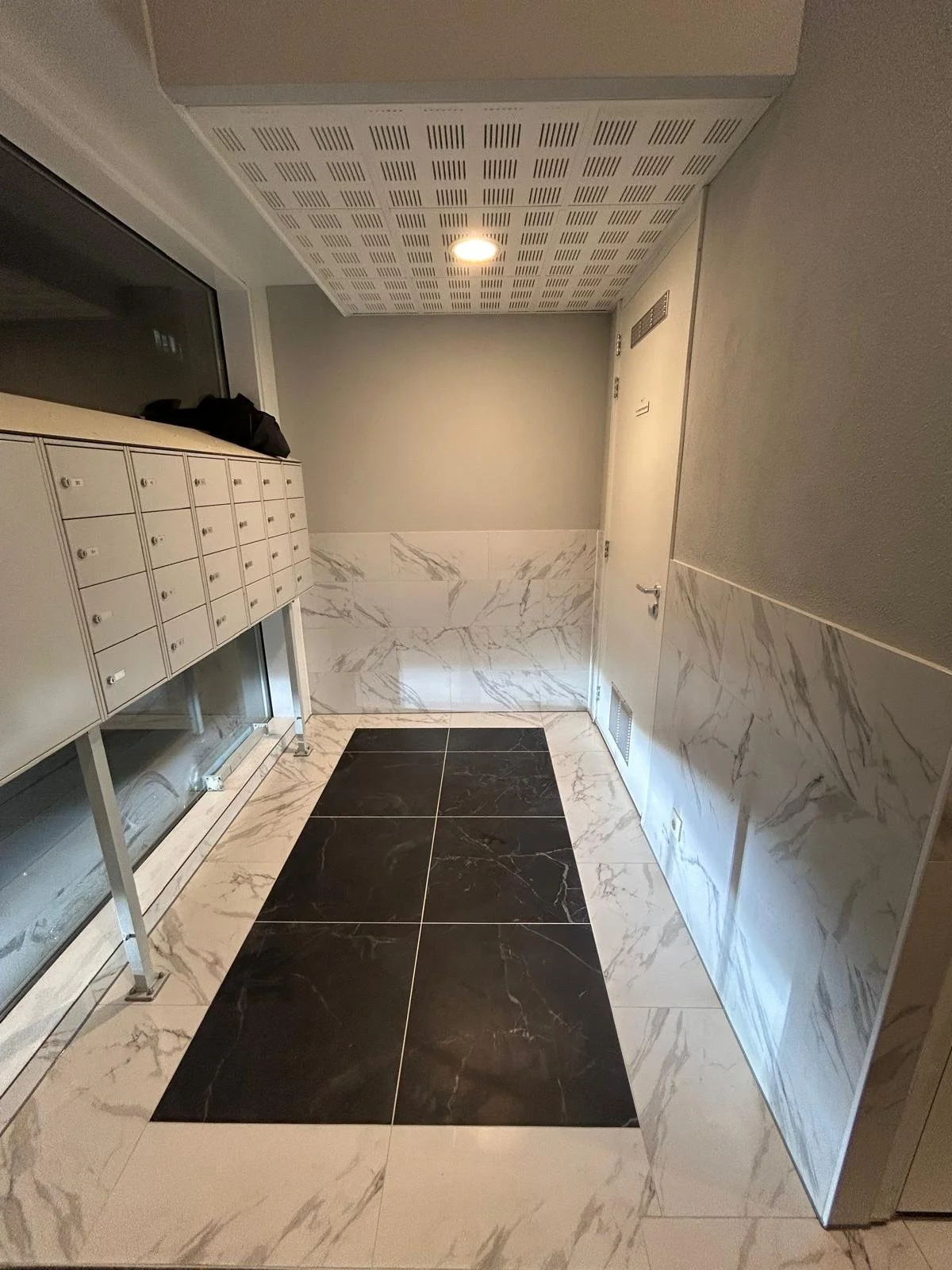Een smalle hal met een wit plafond met ventilatiekuiltjes, beige muren, een witte deur, waarop een kleine ventilatierooster zit, en grijze en witte marmeren wandbekleding. On de vloer liggen grote witte en zwarte marmeren tegels.