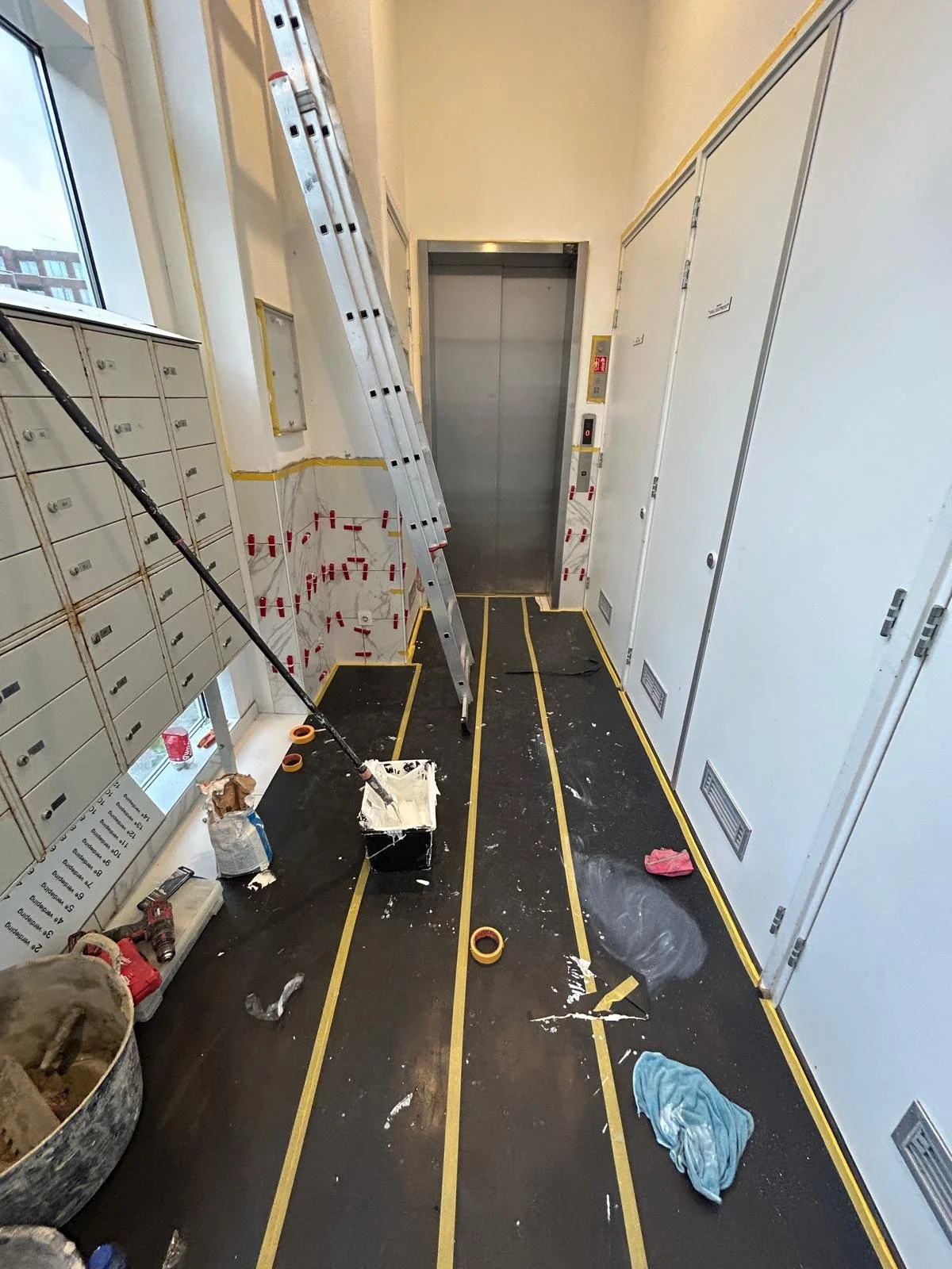 Het interieur van een bouw- of renovatieplek met een lijntape op de vloer, een ladder, verfspatten, verfemmer en bouwmaterialen, en een liftdeur op de achtergrond.