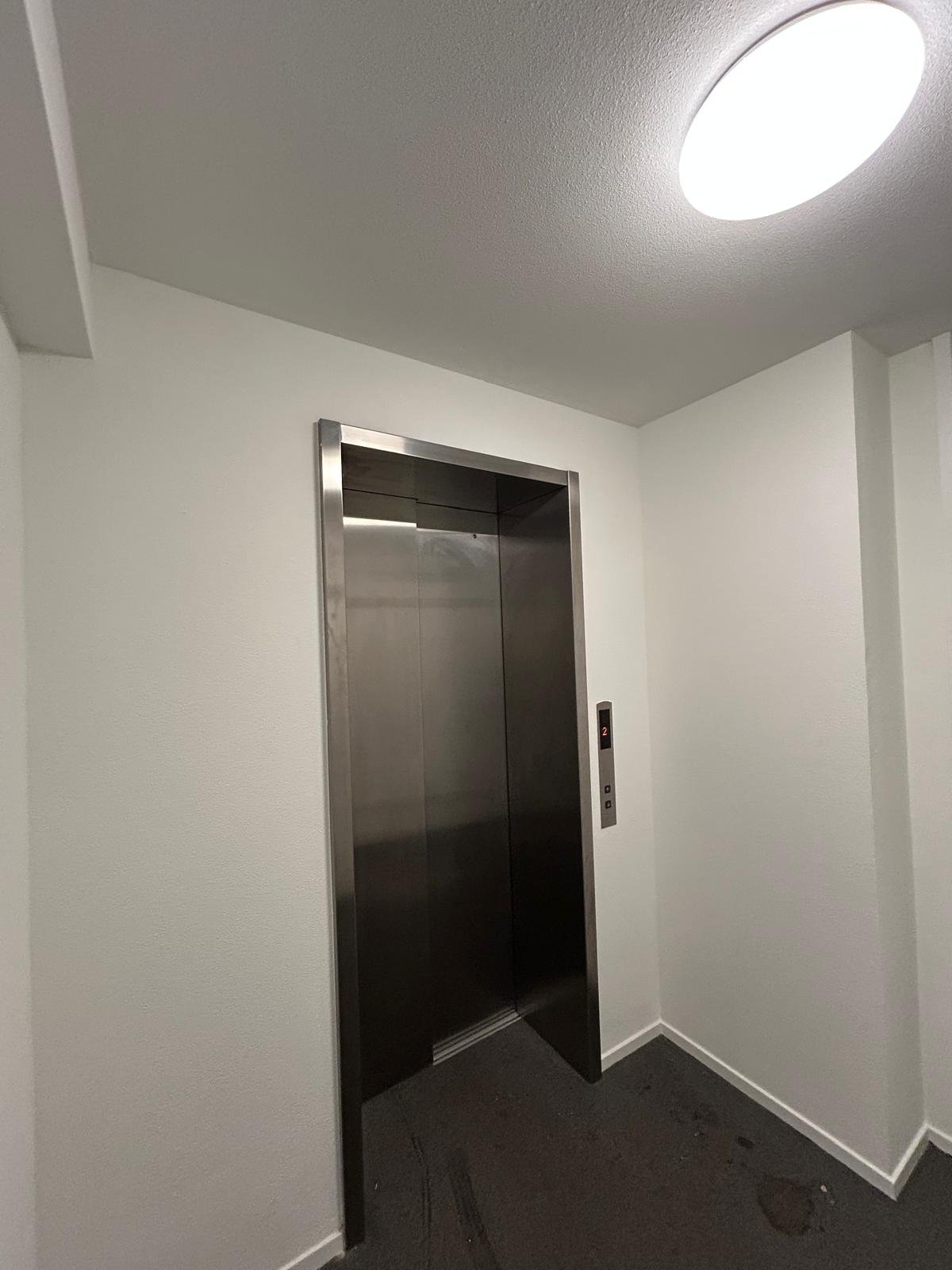 Staalkleurige liftdeur in een witte muur, met een bedieningspaneel aan de rechterkant.