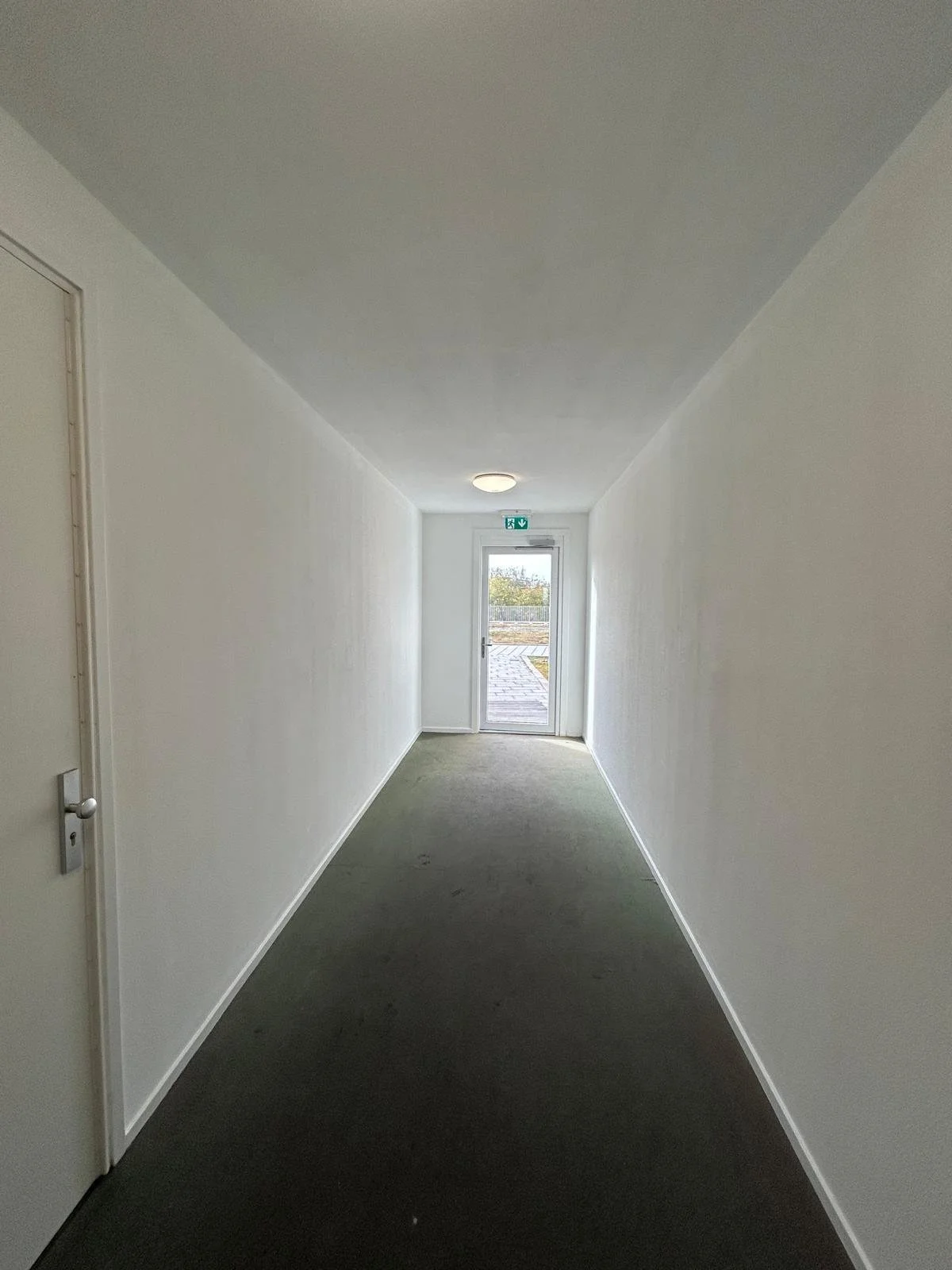 Een smalle, lege wit geschilderde gang met een deur aan de linkerzijde, een plafondlamp en een uitgang met een glazen deur aan het einde, die uitkomt op een terras buiten.
