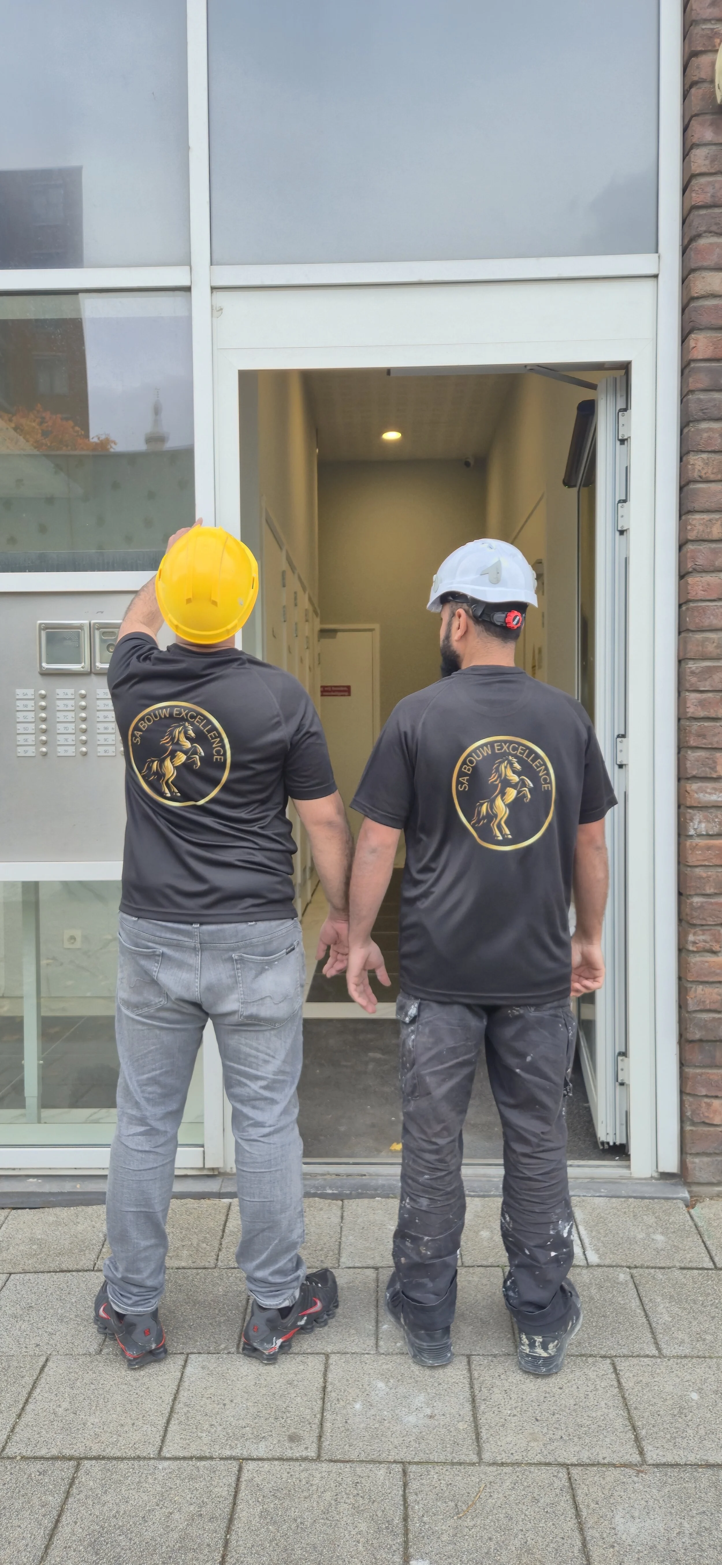 Twee bouwvakkers, één met een geel helm en één met een wit helm, staan voor een buitendeur, kijkend naar binnen. Ze dragen zwarte T-shirts met een gouden logo en de tekst 'SB Bouw Excellence', en werkbroeken. Ze lijken overleg te plegen tijdens bouw- of renovatiewerkzaamheden.