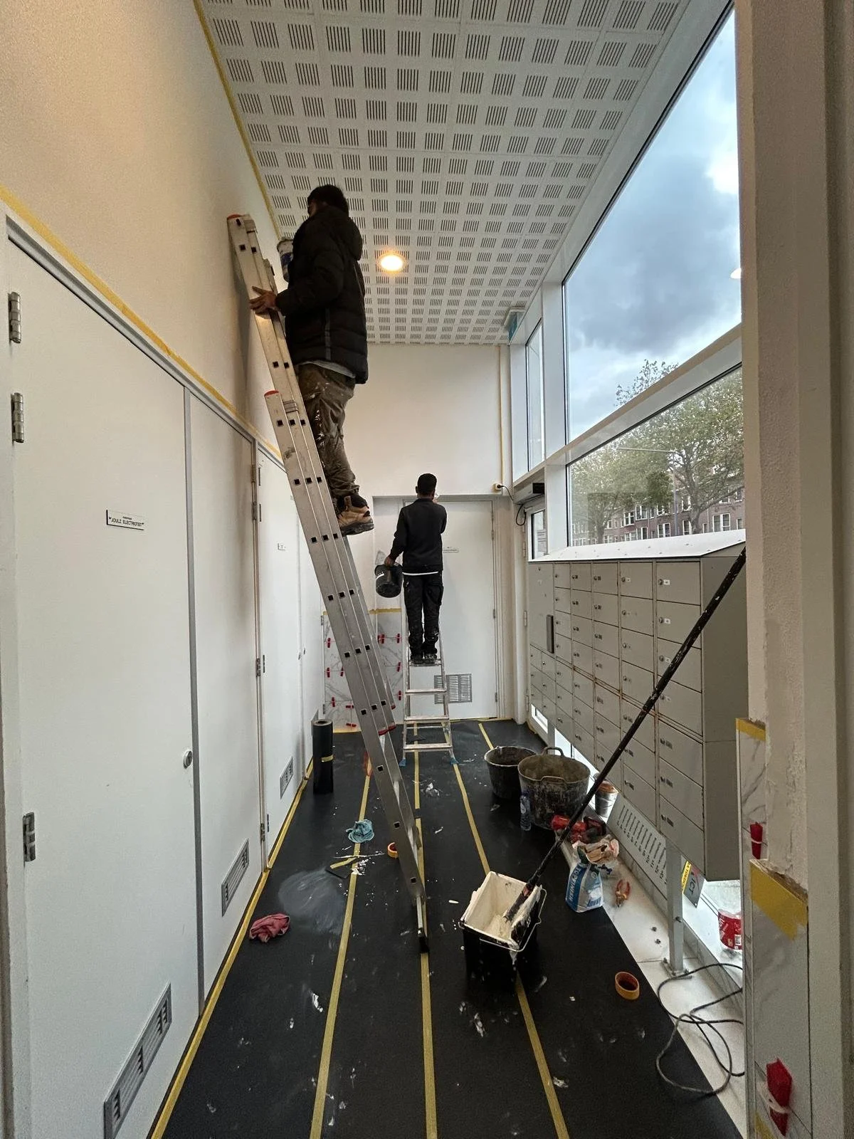 Twee arbeiders installeren of onderhouden het plafonsysteem in een hal met grote ramen, er is bouw- of onderhoudsmaterieel en vuil op de vloer.