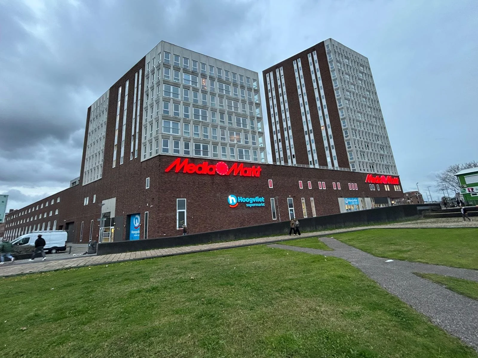 Groot modern gebouw met mediastore en supermarkt, groene grasveld vooraan, bewolkte hemel