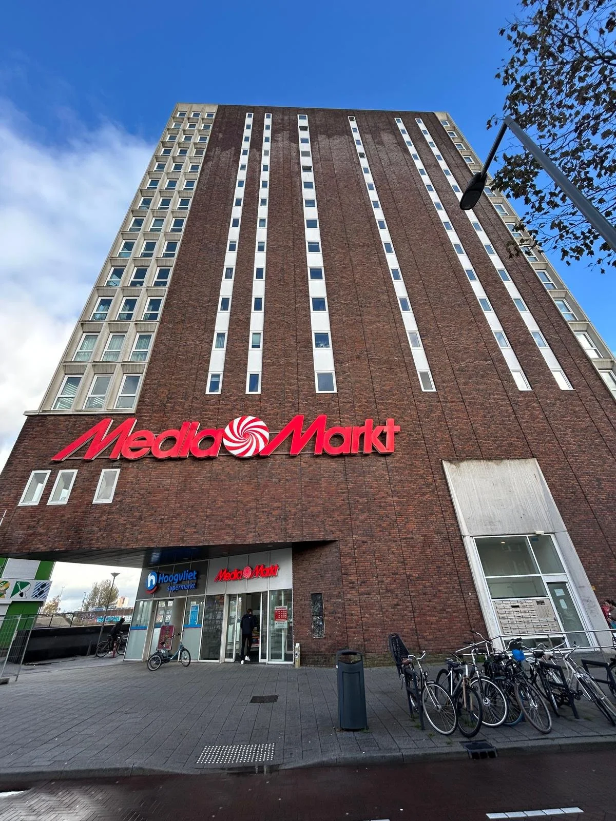 Uitzicht van onderaf op een hoog gebouw met een Media Markt winkel op de begane grond, fietsen geparkeerd voor de ingang en een blauwe lucht met enkele wolken erboven.