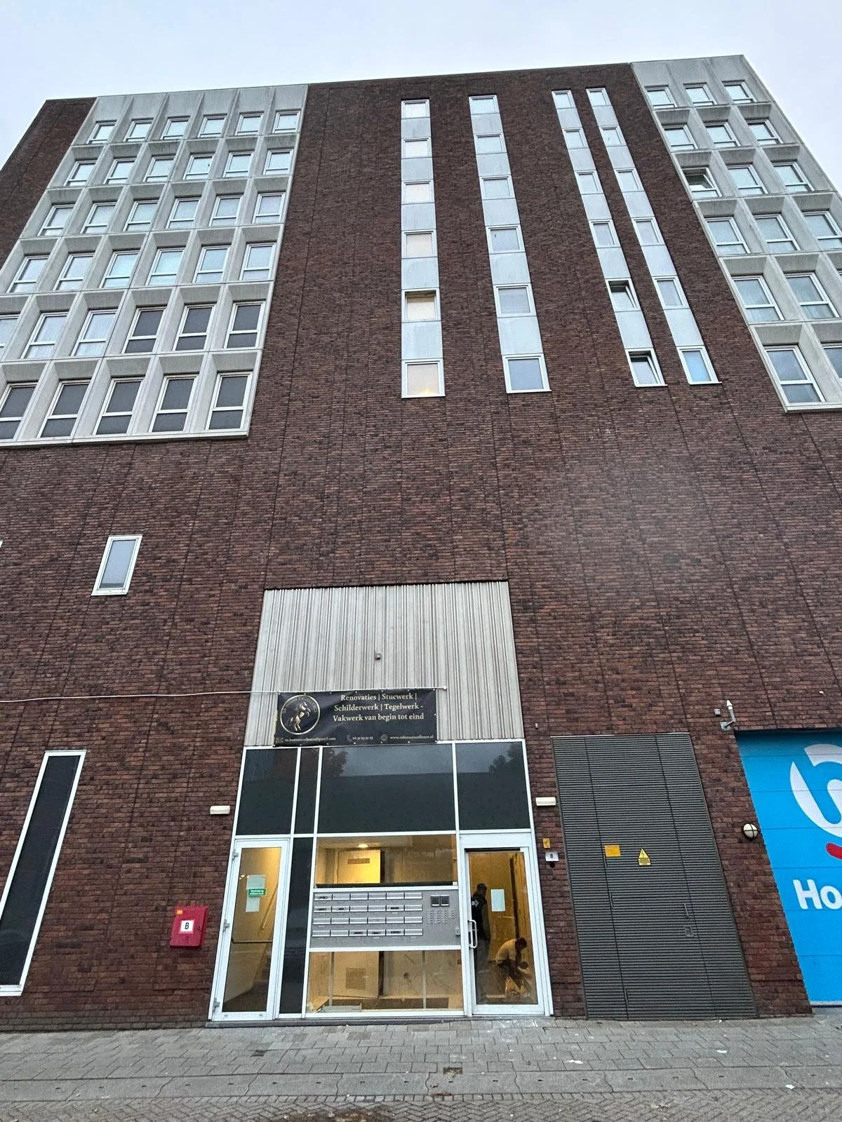 Een hoog appartementencomplex met een rode bakstenen gevel en meerdere rijen ramen. De ingang heeft een glazen deur en een aantal brievenbussen. Een persoon staat binnen.