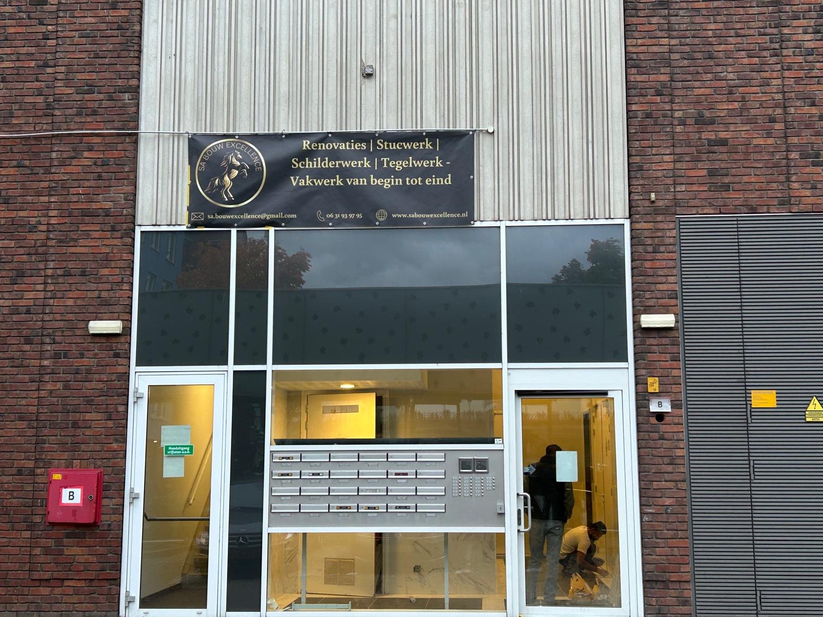 De gevel van een gebouw met een glasdeuren ingang, een bord met bedrijfsnaam singel en contactgegevens, en enkele mensen binnen die werken.