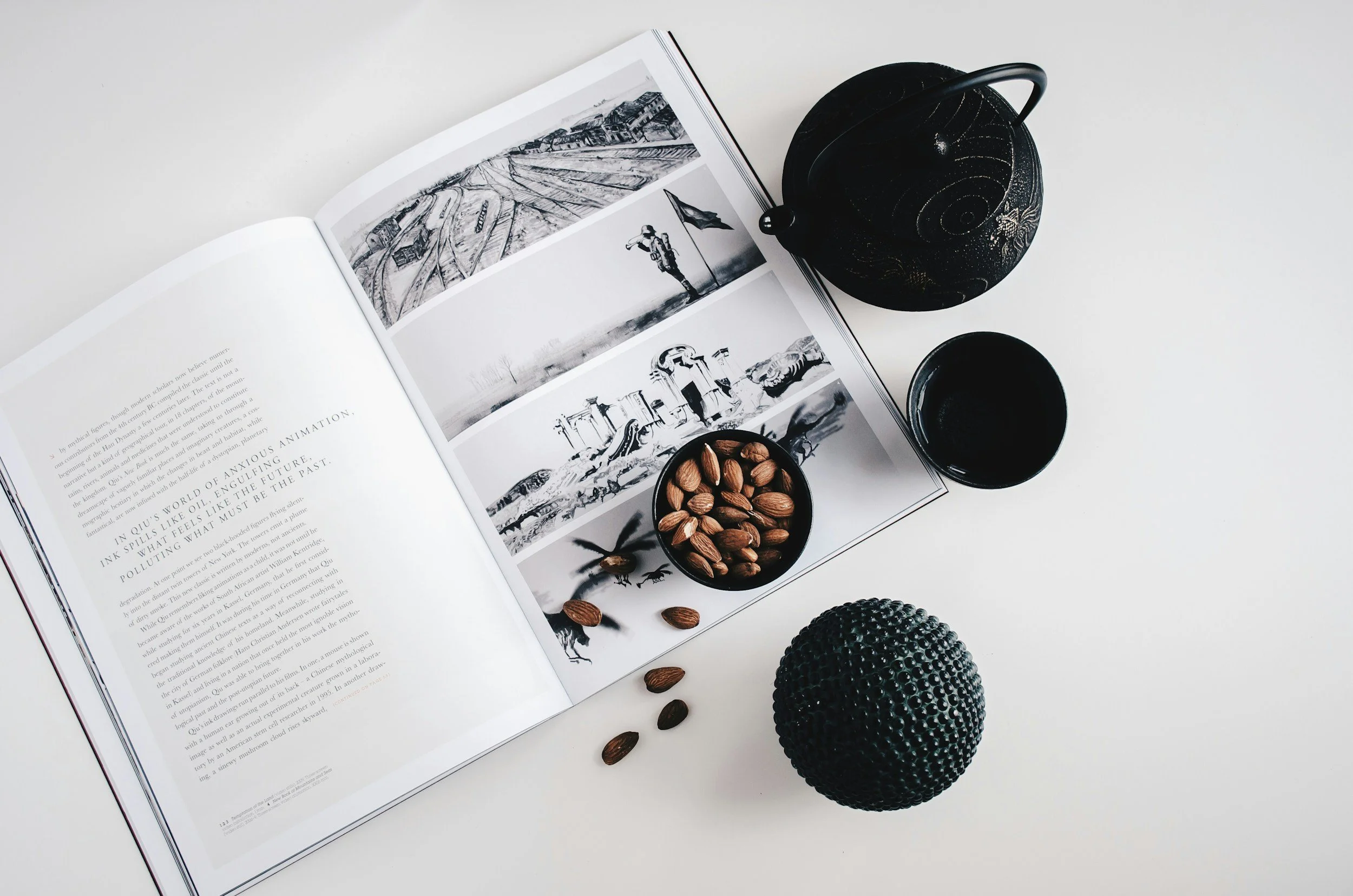 Open magazine with black and white photographs, black decorative item, drinking glass, bowl of almonds, and textured black spherical object on a white surface