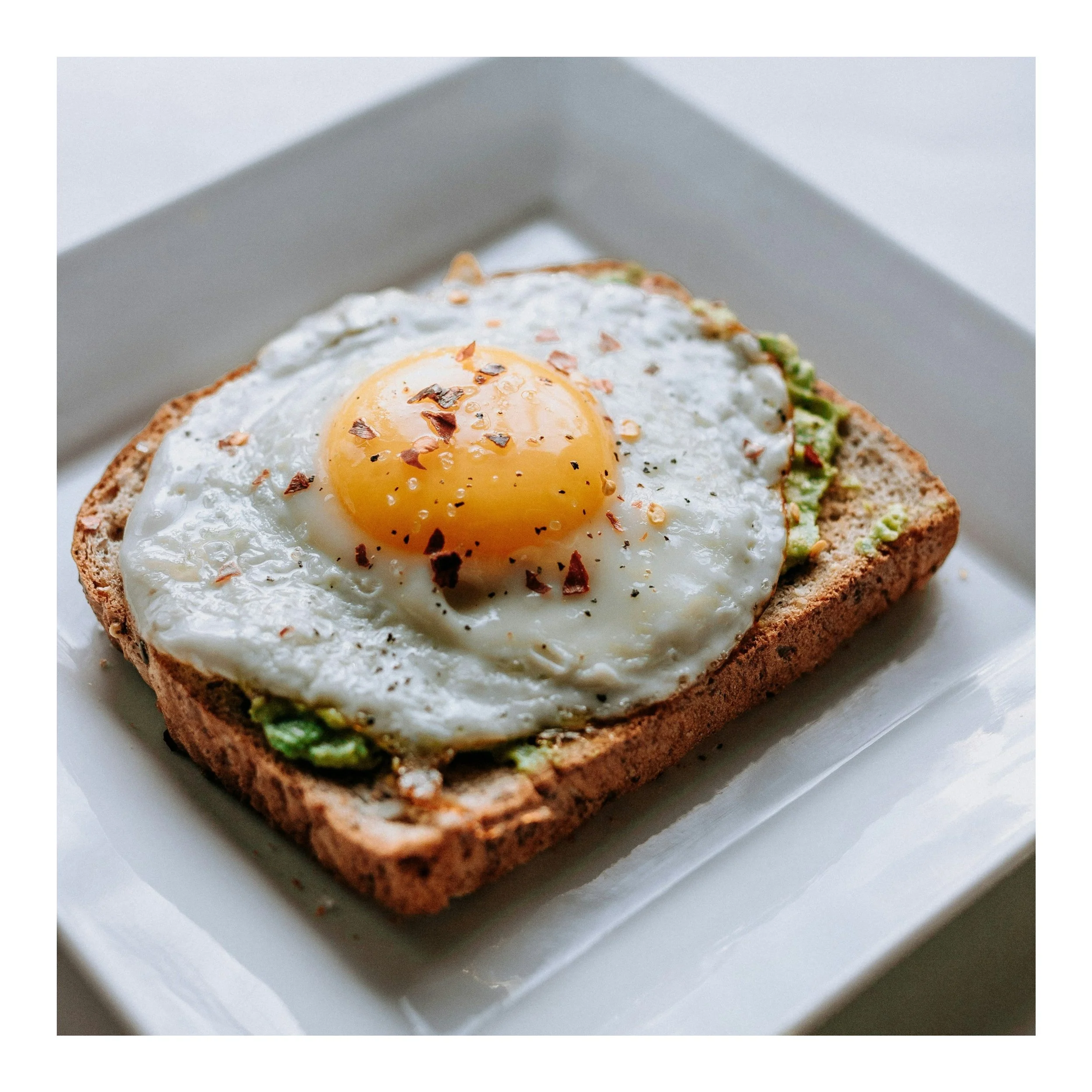 Fried egg on toasted whole grain bread with avocado spread, sprinkled with chili flakes on white plate.