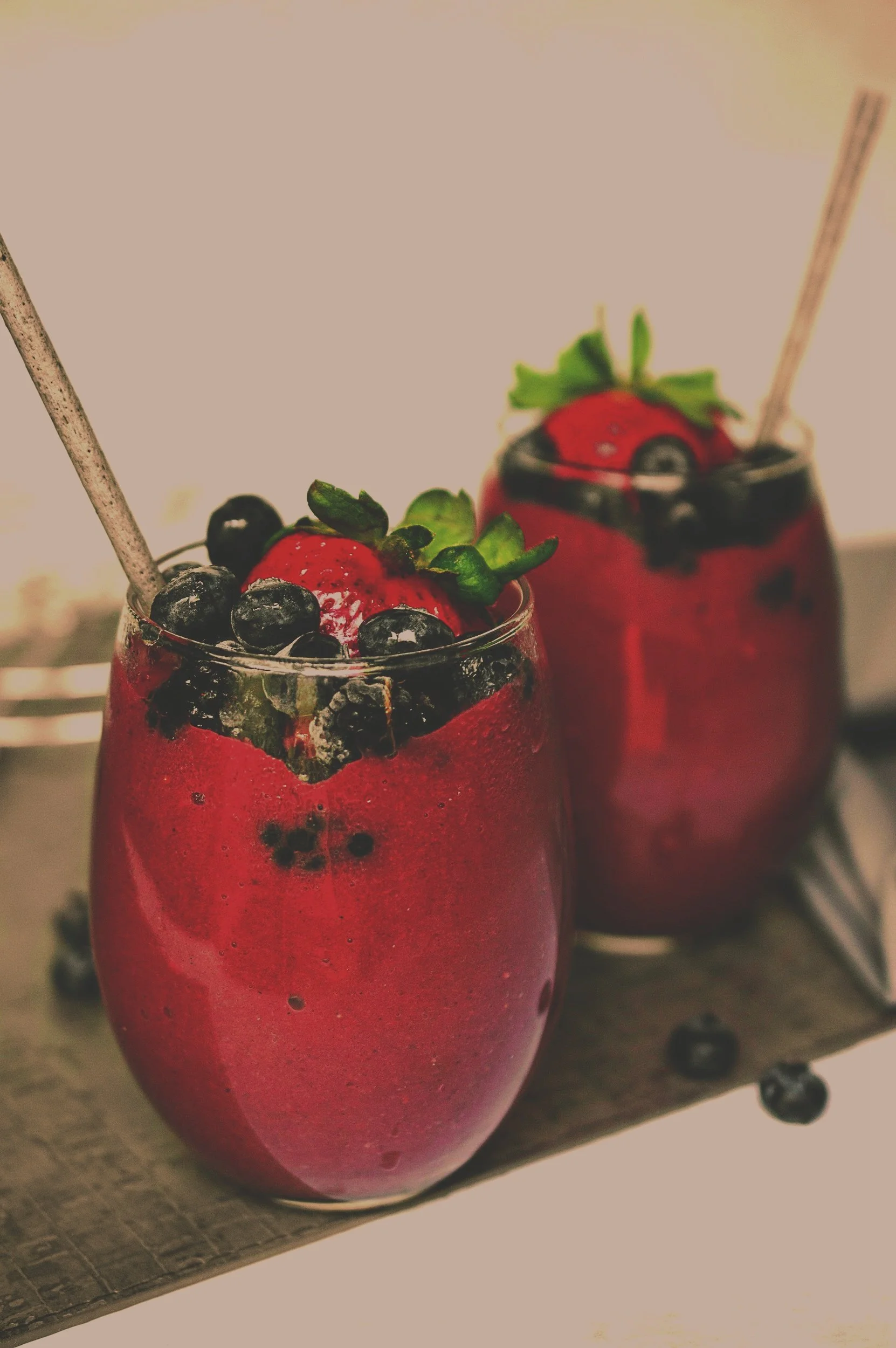 Two glasses of blueberry strawberry smoothies garnished with fresh strawberries and blueberries, with wooden spoons in each, placed on a wooden surface.