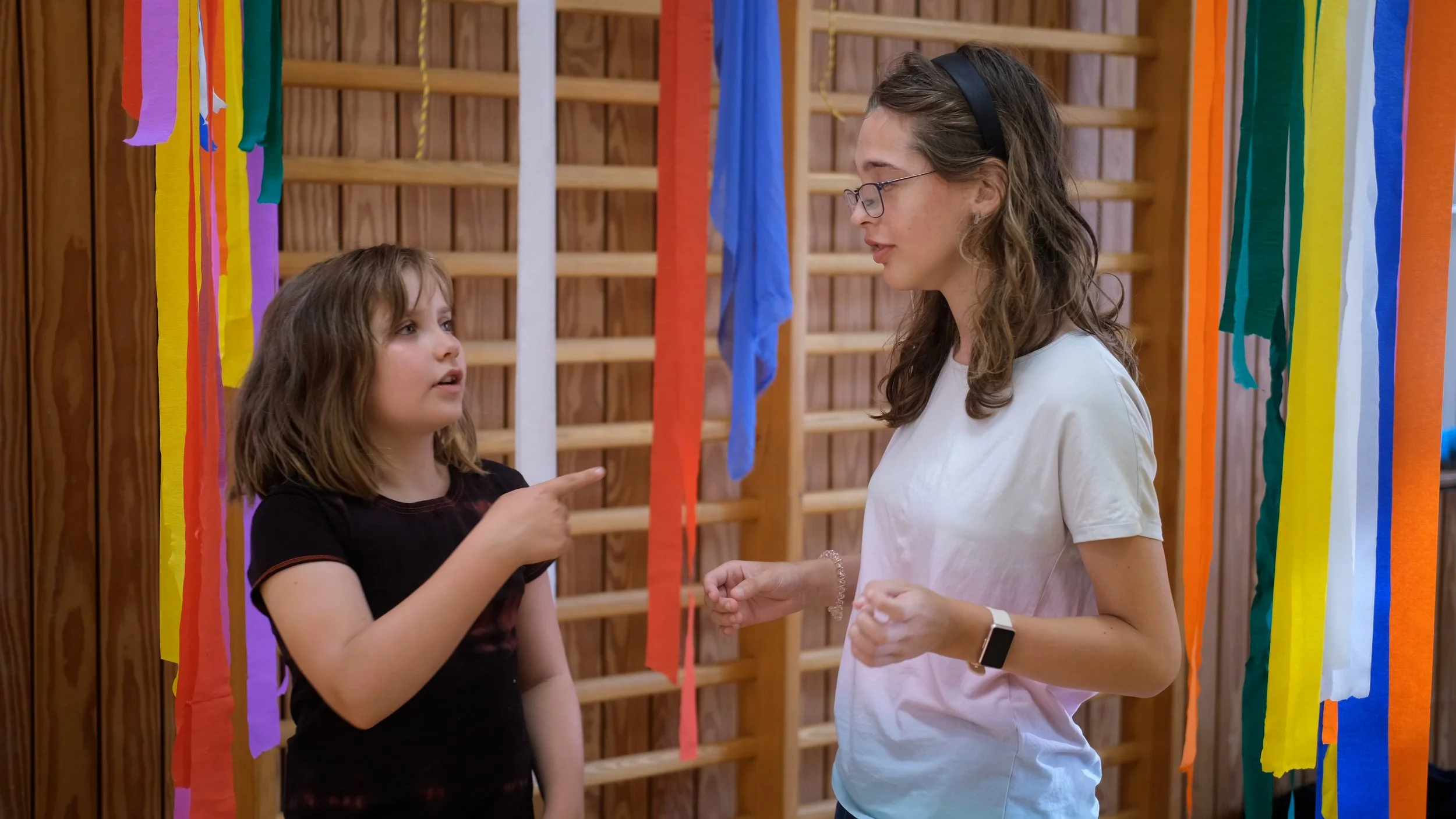 Zwei Kinder, Tjade, 10 Jahre und Timasia, 13 Jahre stehen sich in einer Sporthalle gegenüber. Tjade zeigt auf Timasia. Um sie herum hängen bunte Kreppbänder.