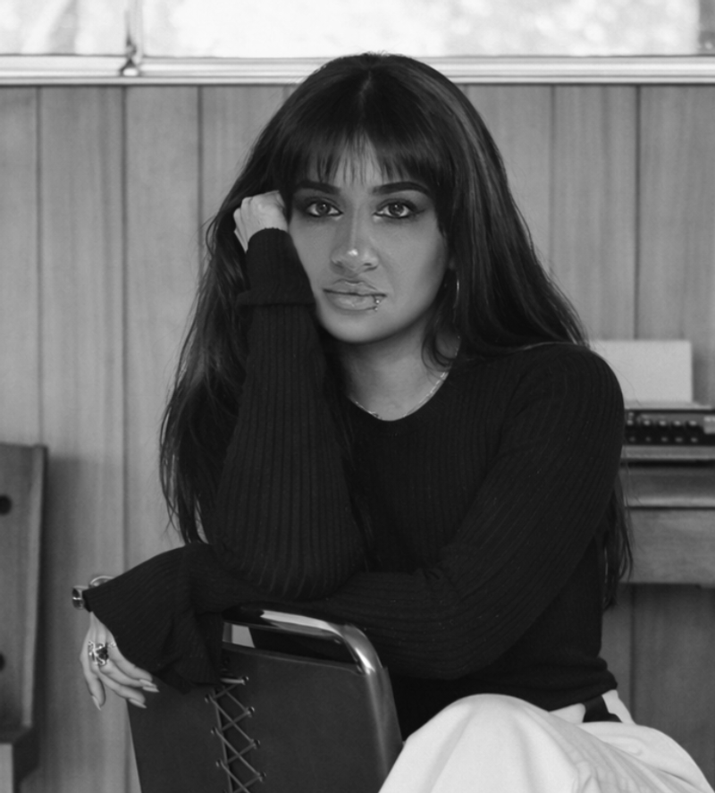 Black and white photo of a young woman with long dark hair, sitting on a chair in a room with wood-paneled walls, resting her chin on her hand, wearing a long-sleeve black top and light-colored pants.