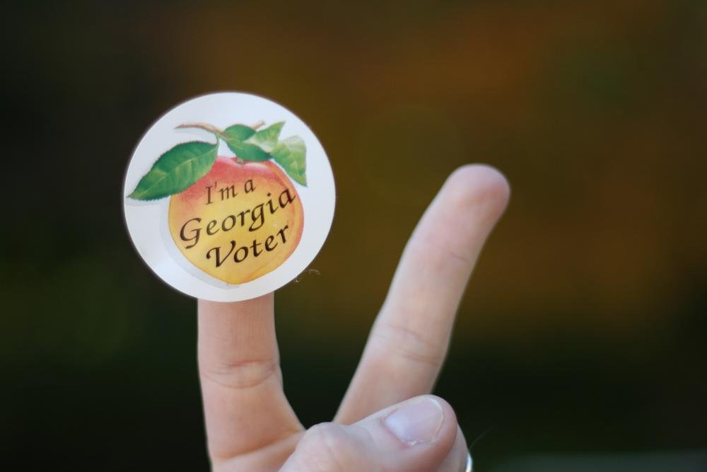 A hand holding a small sticker with a voting logo and the word "VOTED" in front of a dark background.