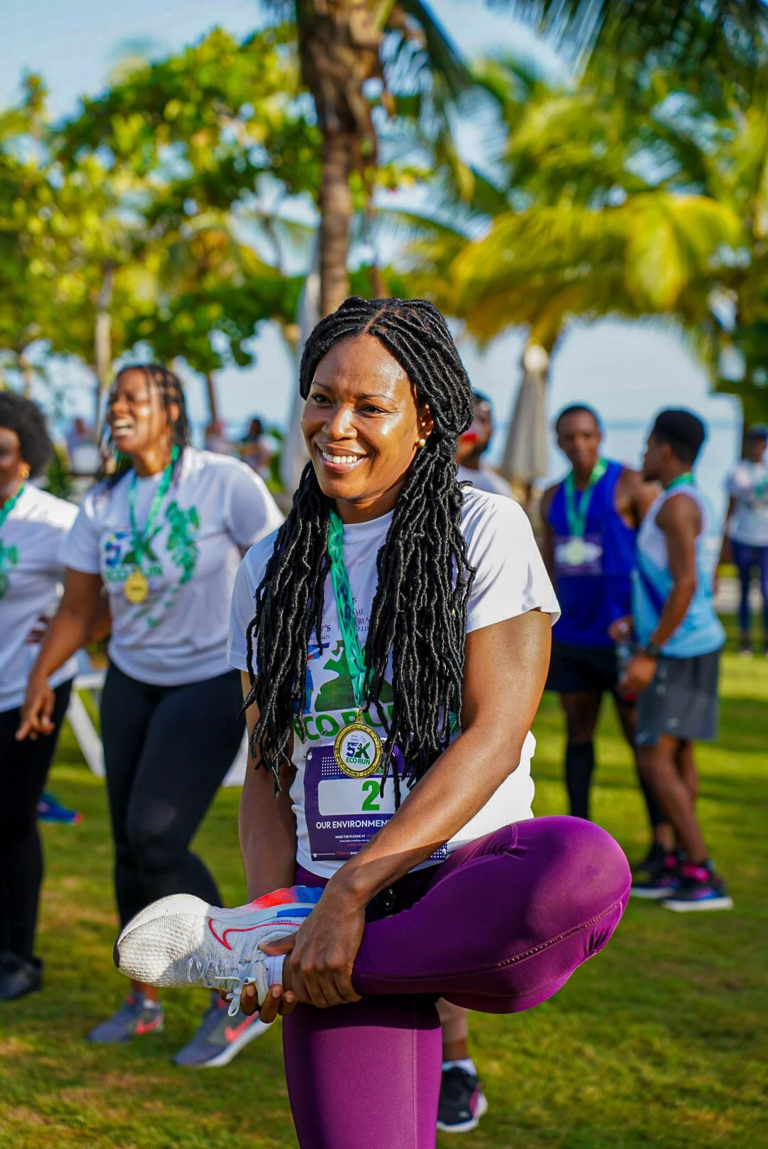 Woman stretching before a 5K run outdoors, wearing a white t-shirt with event logo, purple leggings, holding her leg up. Other participants in the background with trees and clear sky.