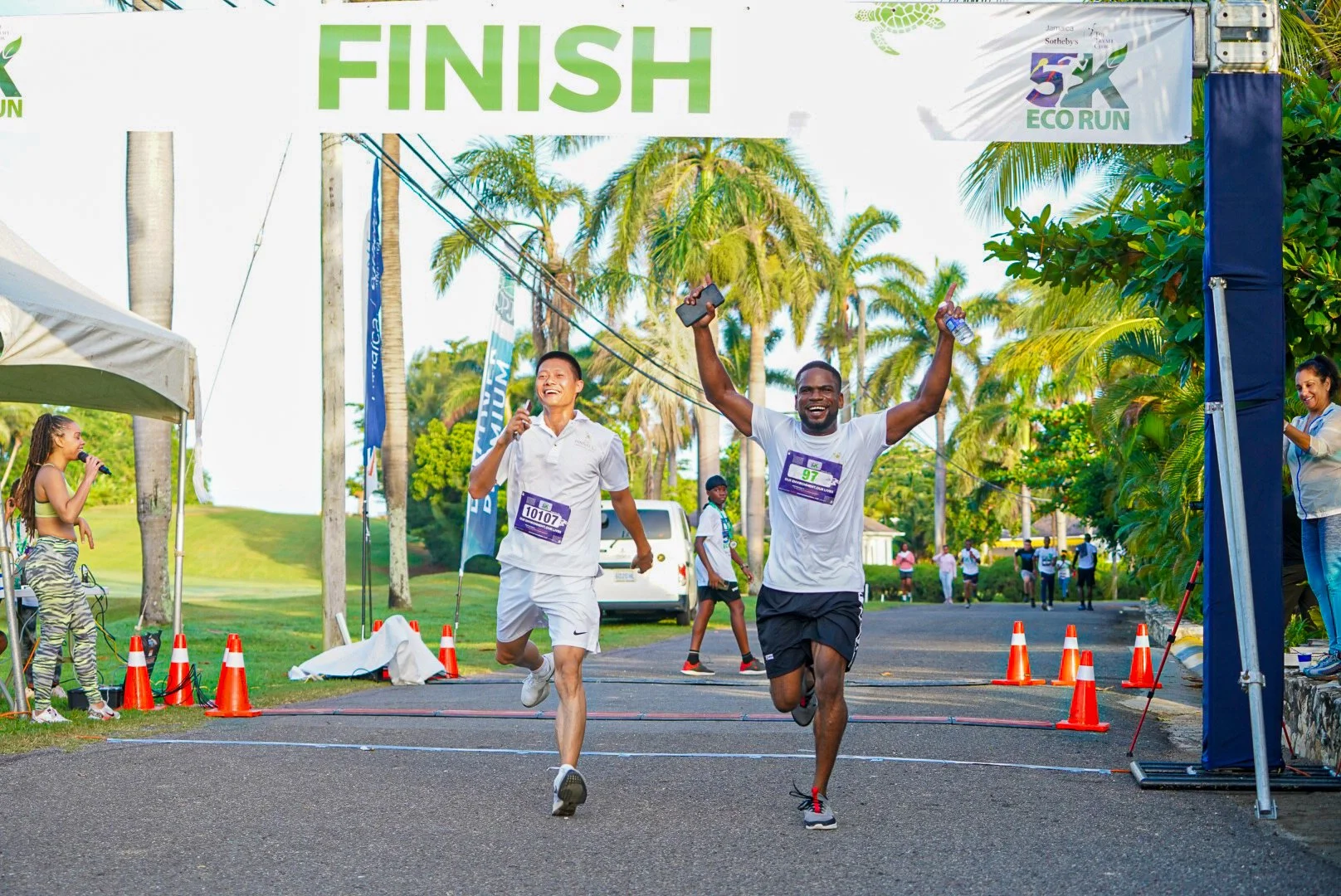 Two male runners celebrating as they cross the finish line of a 5K eco run, surrounded by palm trees.