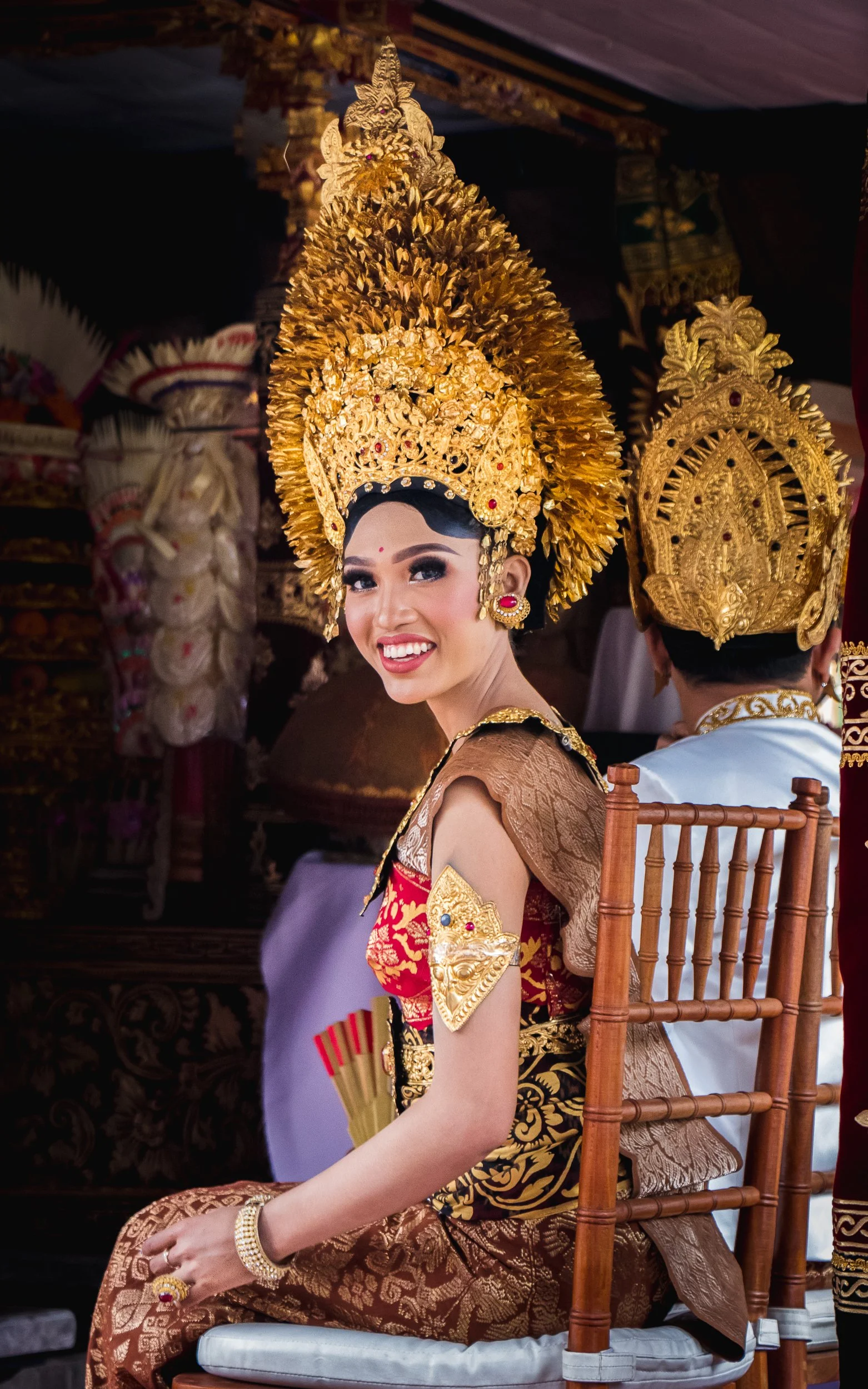 A woman dressed in traditional Indonesian attire with elaborate gold headdress, smiling while seated on a wooden chair during a cultural event.