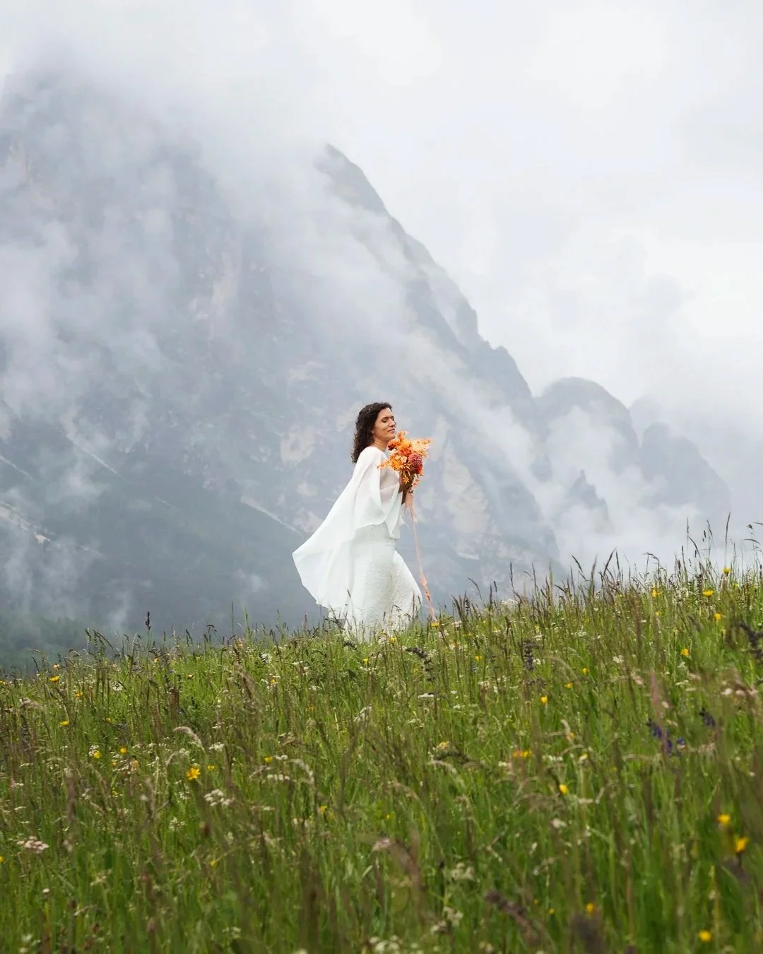 Woman in white dress holding a bouquet of orange and yellow flowers standing in a green meadow with tall grass and wildflowers, mountain range with clouds in the background.