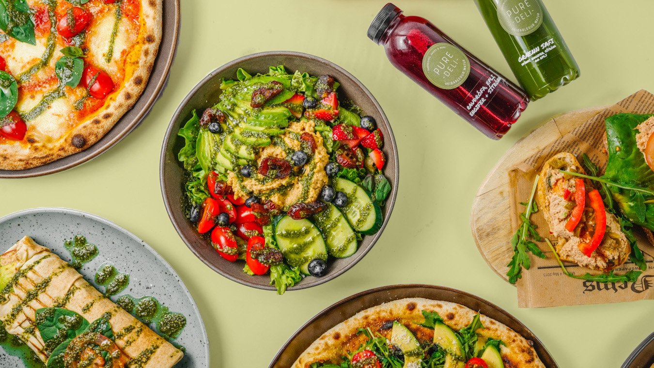 Assorted food items including two pizzas, a salad with strawberries and blueberries, a wrap, and two bottles of juice on a light green surface.
