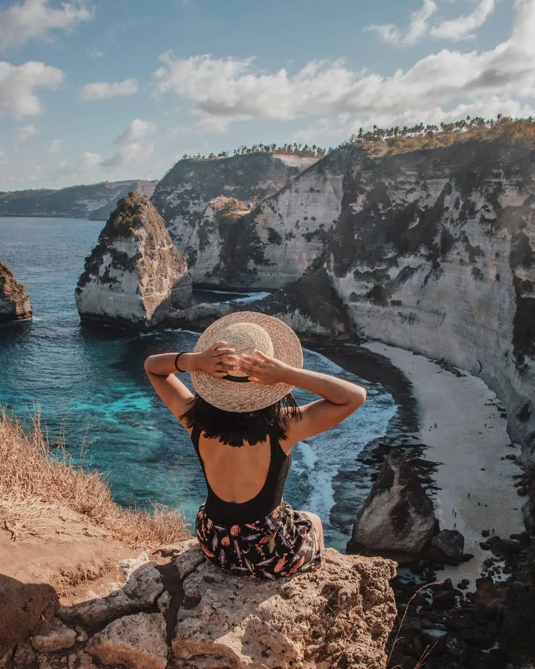 A woman sitting on a rocky cliff, wearing a large sun hat and a black top, overlooks a scenic coastal landscape with white cliffs, rock formations, and a blue ocean under a partly cloudy sky.