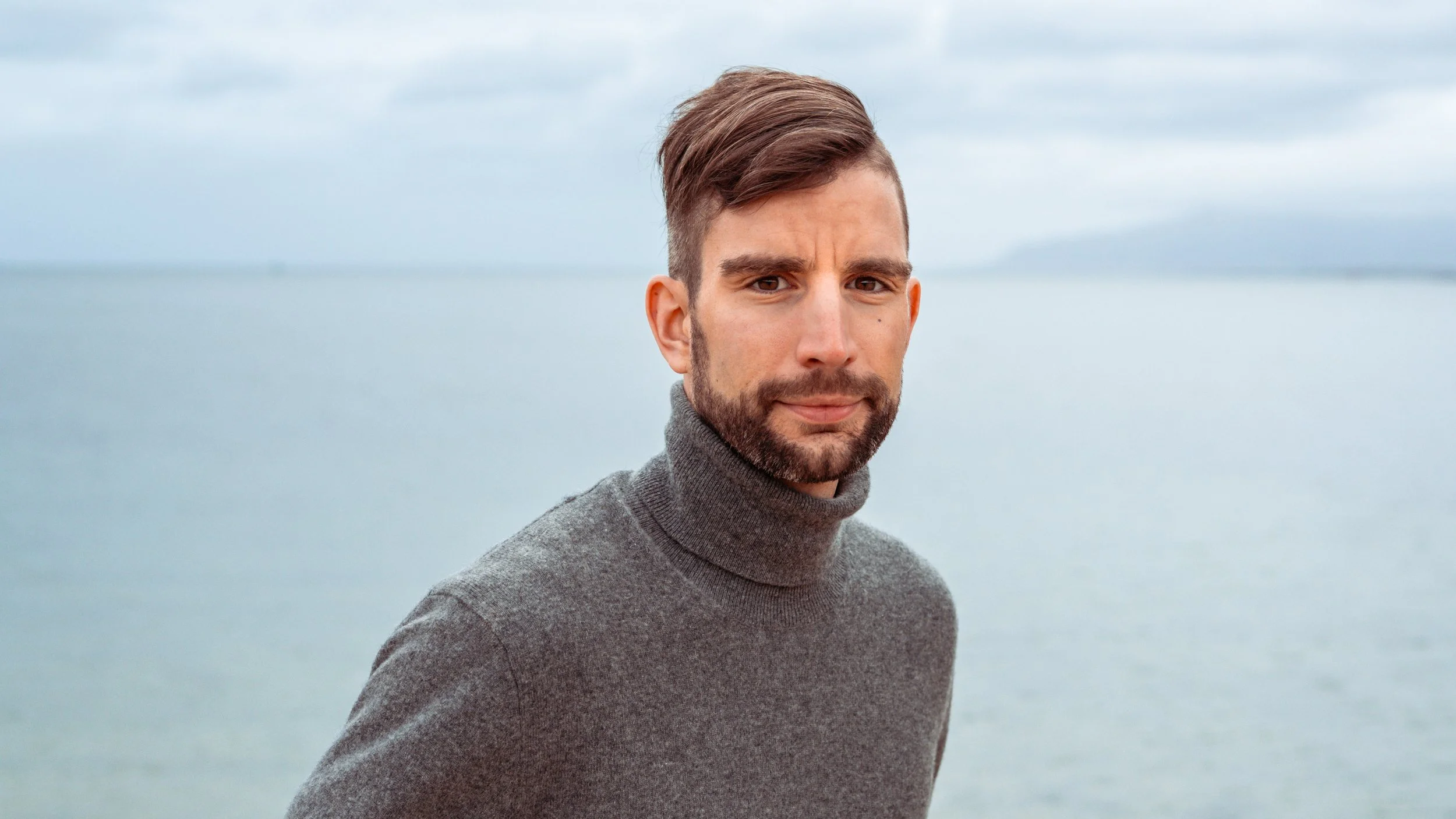Man with brown hair and beard in a gray turtleneck sweater standing near water with cloudy sky in background.