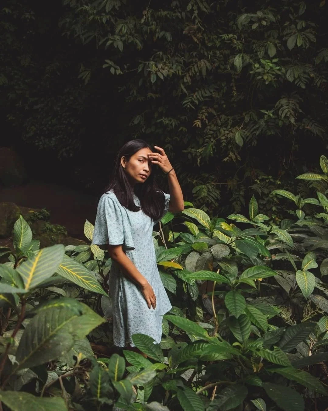 A woman with long dark hair standing among lush green plants in a dense forest at night, wearing a light blue dress, touching her forehead with one hand, looking contemplative.