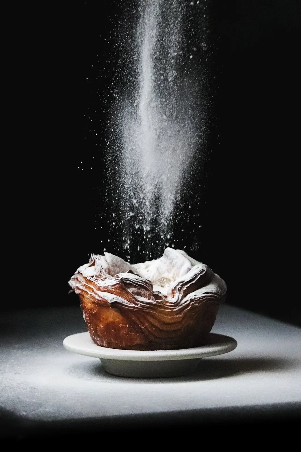A dessert topped with whipped cream on a white plate, with powdered sugar being dusted from above against a dark background.