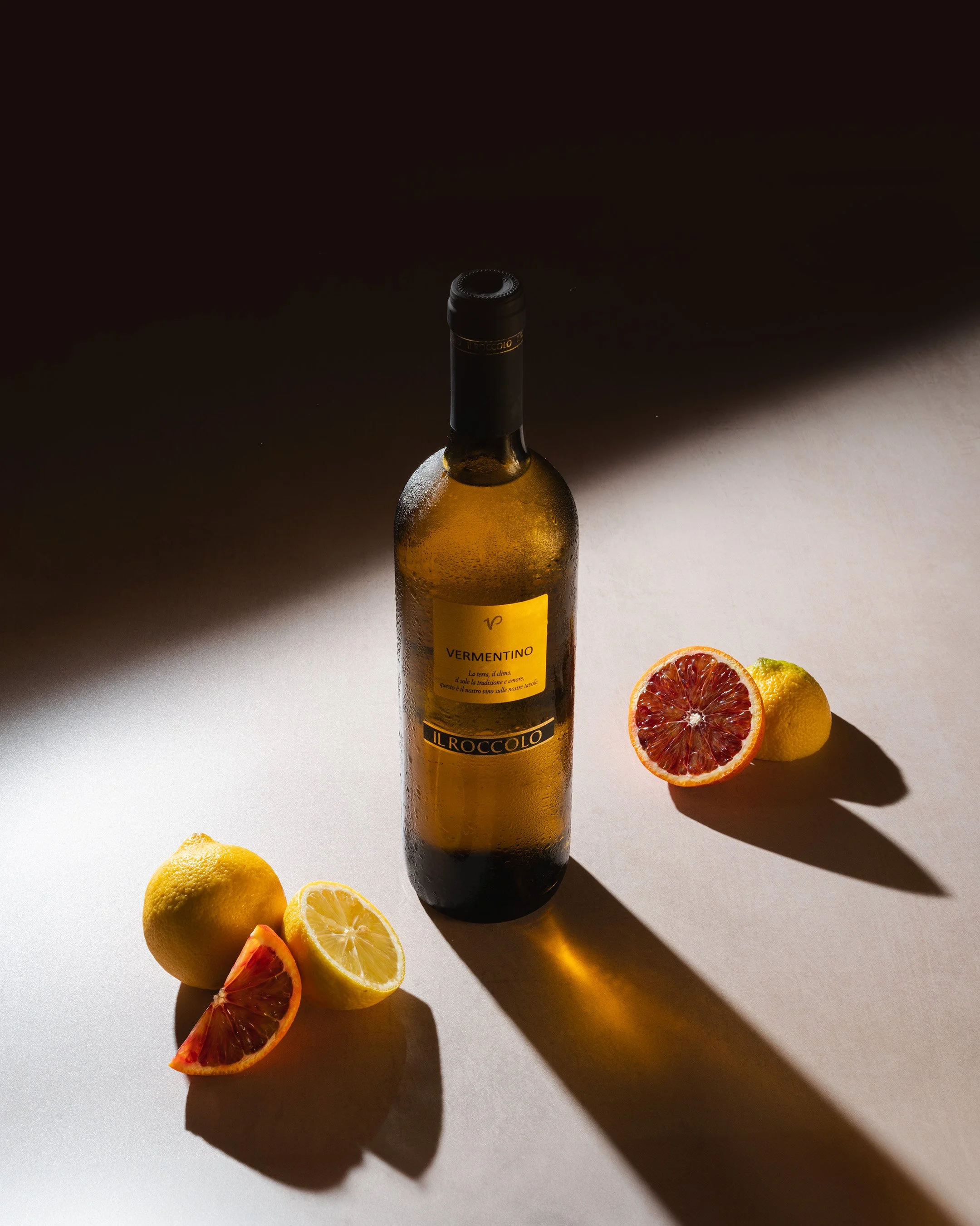 A bottle of white wine labeled 'Vermentino' on a light gray surface, with lemons and blood oranges arranged around it, casting shadows in a dimly lit setting.