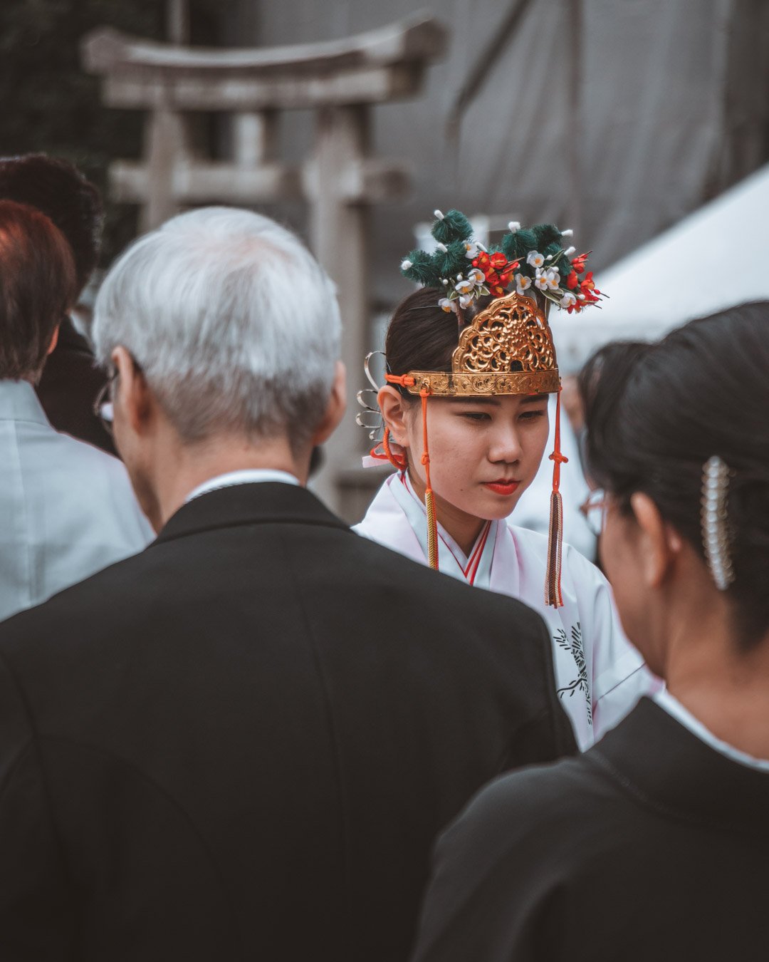 A young woman dressed in traditional Korean wedding attire, including a white hanbok and ornate headpiece, participating in a cultural wedding ceremony surrounded by people.
