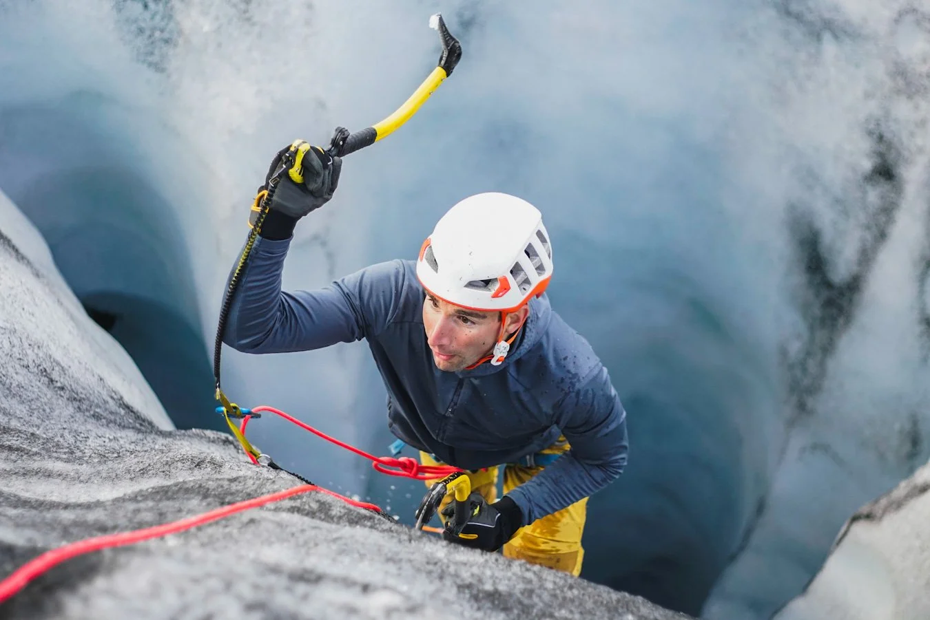 A rock climber wearing a white helmet and yellow climbing pants ascending a steep, rocky cliff.
