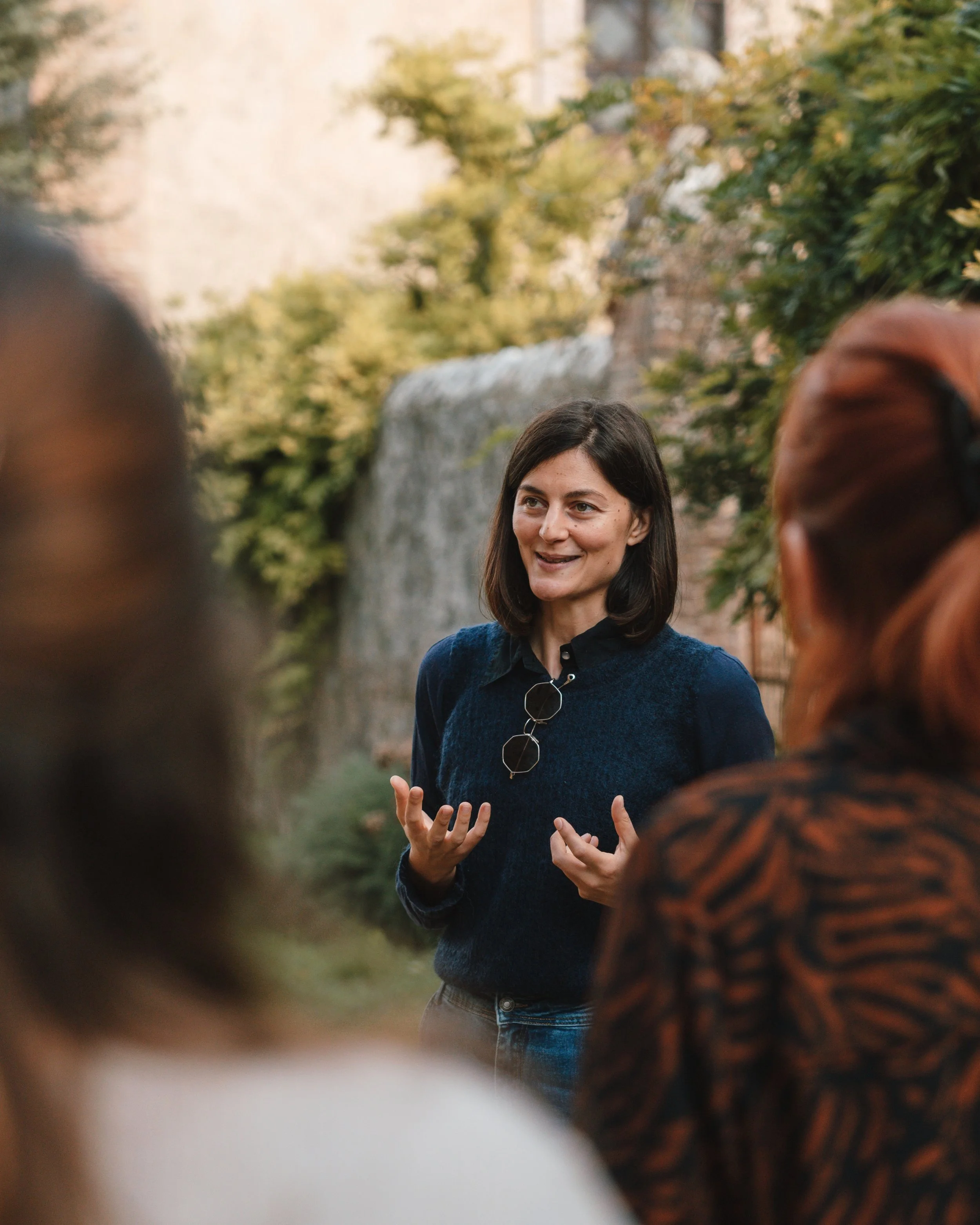 A woman in a navy sweater speaking to a group outdoors with a waterfall and trees in the background.