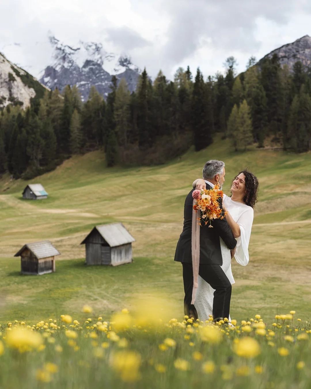 A couple in wedding attire dancing in a grassy field with yellow flowers, surrounded by small wooden cabins and a backdrop of mountains and dense forest.
