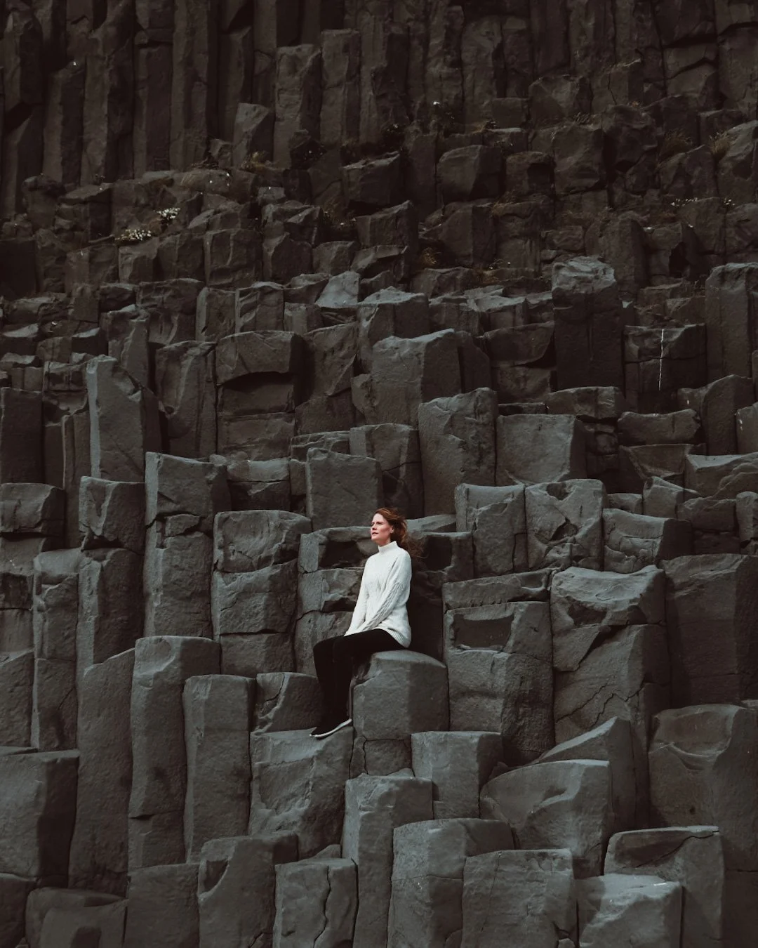 A woman in a white sweater sitting on rocks among towering basalt columns in a natural, rugged landscape.