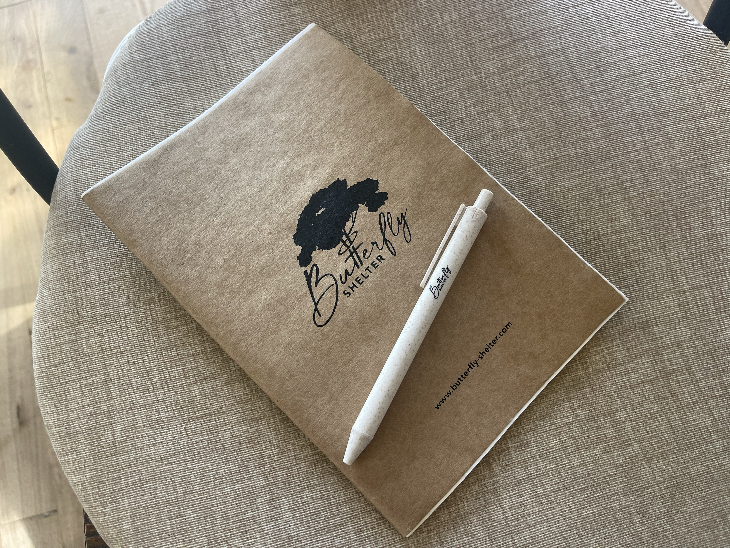 Un carnet en papier kraft avec un logo d'un arbre et le texte « Buttery Shelter » posé sur une table en tissu beige, à côté d'un stylo blanc.