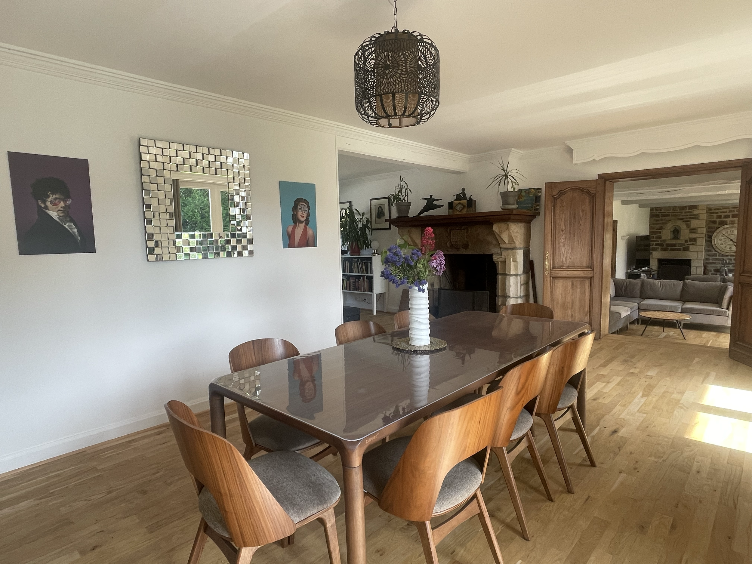 Une salle à manger avec une table en bois entourée de chaises en bois avec coussins gris. Au centre de la table, une vase blanche avec des fleurs violettes et roses. Sur le mur blanc, il y a deux œuvres d'art, une avec un portrait sérieux d'une perso