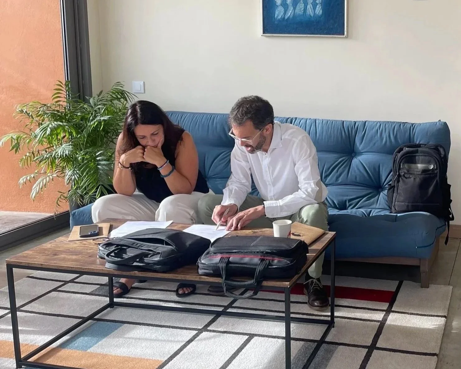 Deux personnes assises sur un canapé en train de regarder des documents sur une table en bois dans un salon, avec un sac à dos noir posé à côté d'eux.