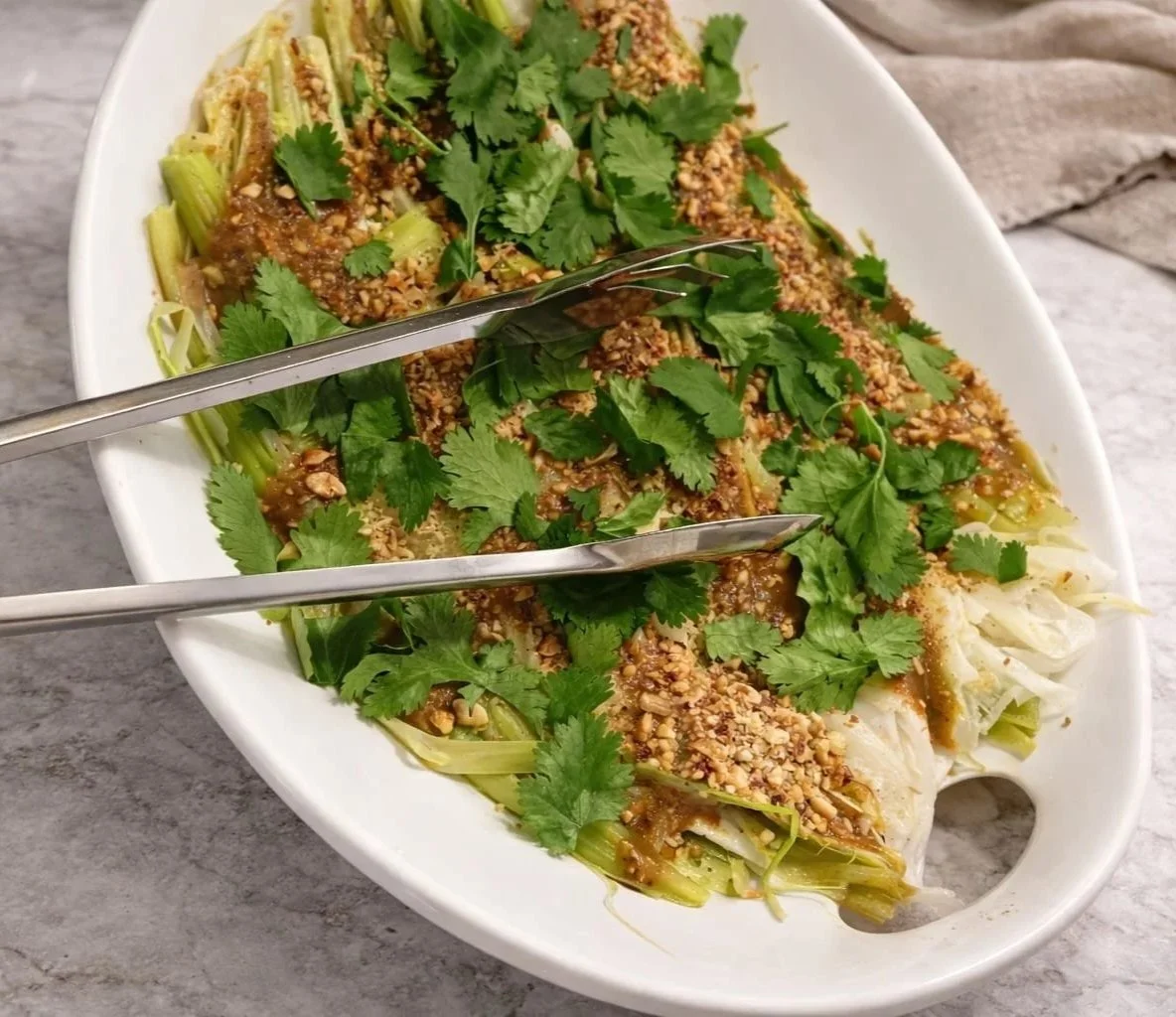 Une salade dans un plat blanc, garnie de coriandre, de graines concassées et de sauce, avec des pinces pour servir.