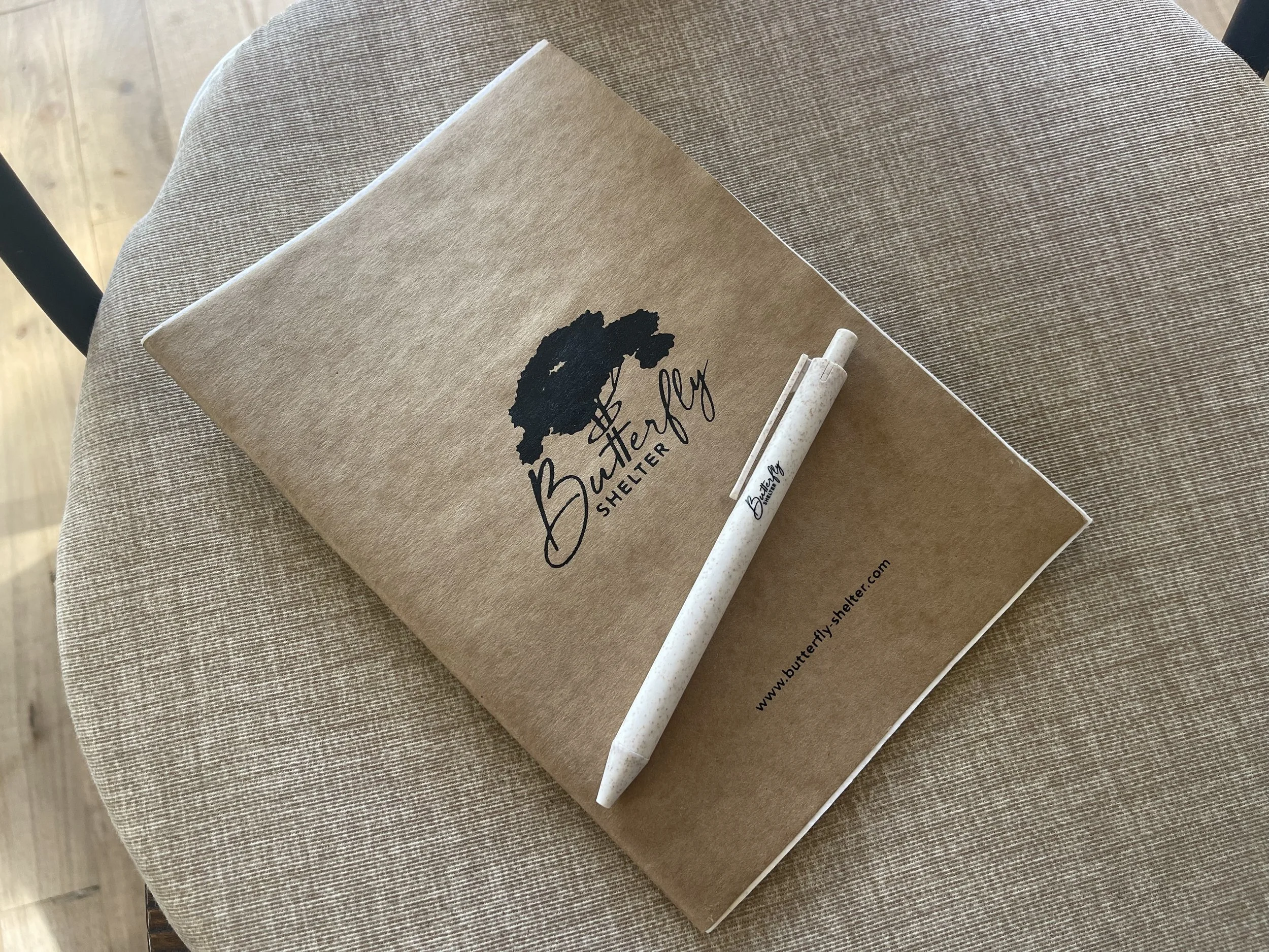 Un carnet de notes avec une illustration d'un arbre et le texte « Butterly Shelter » posé sur une table en tissu beige avec un stylo blanc à côté.