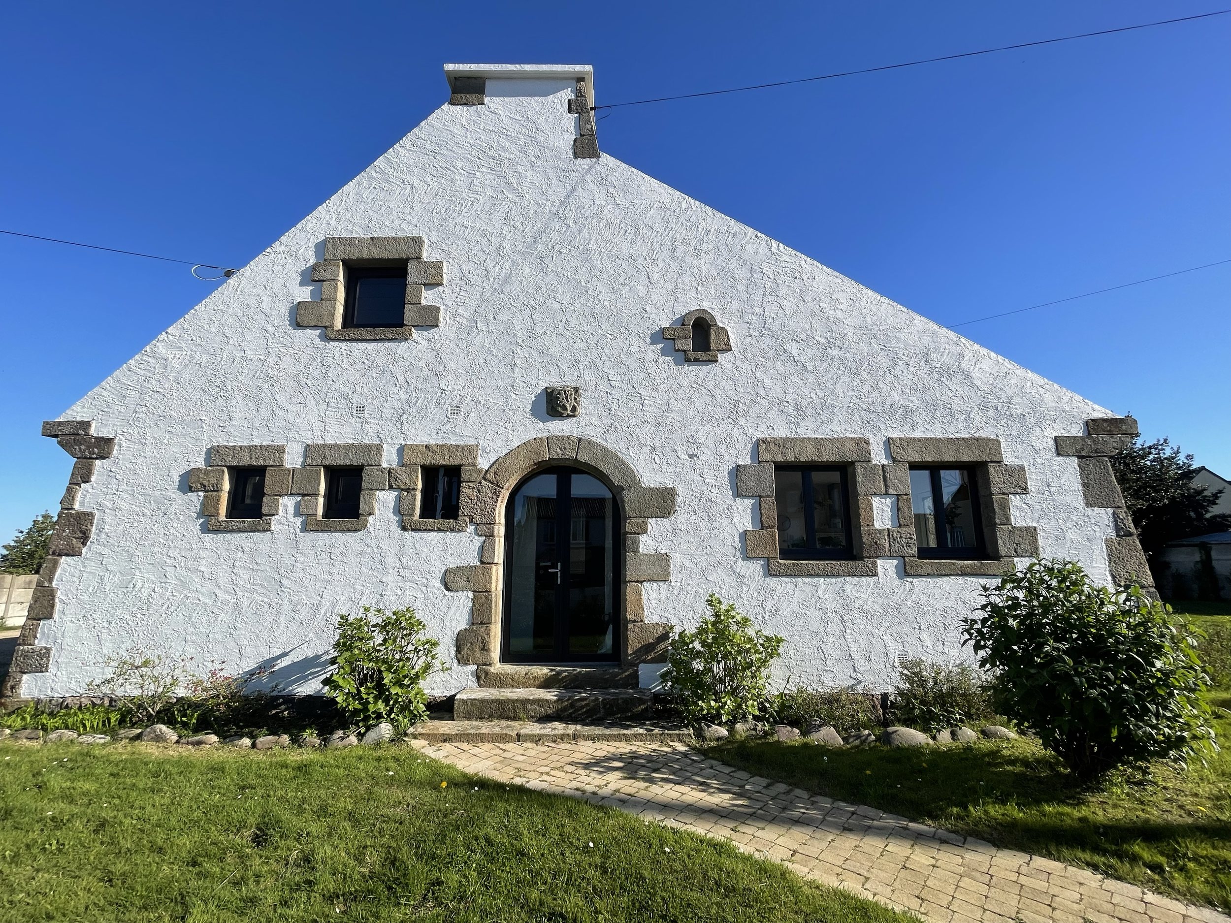 Une maison en pierre blanche avec des fenêtres encadrées de pierre et un porche, entourée d'un jardin vert avec un chemin pavé, sous un ciel bleu clair.