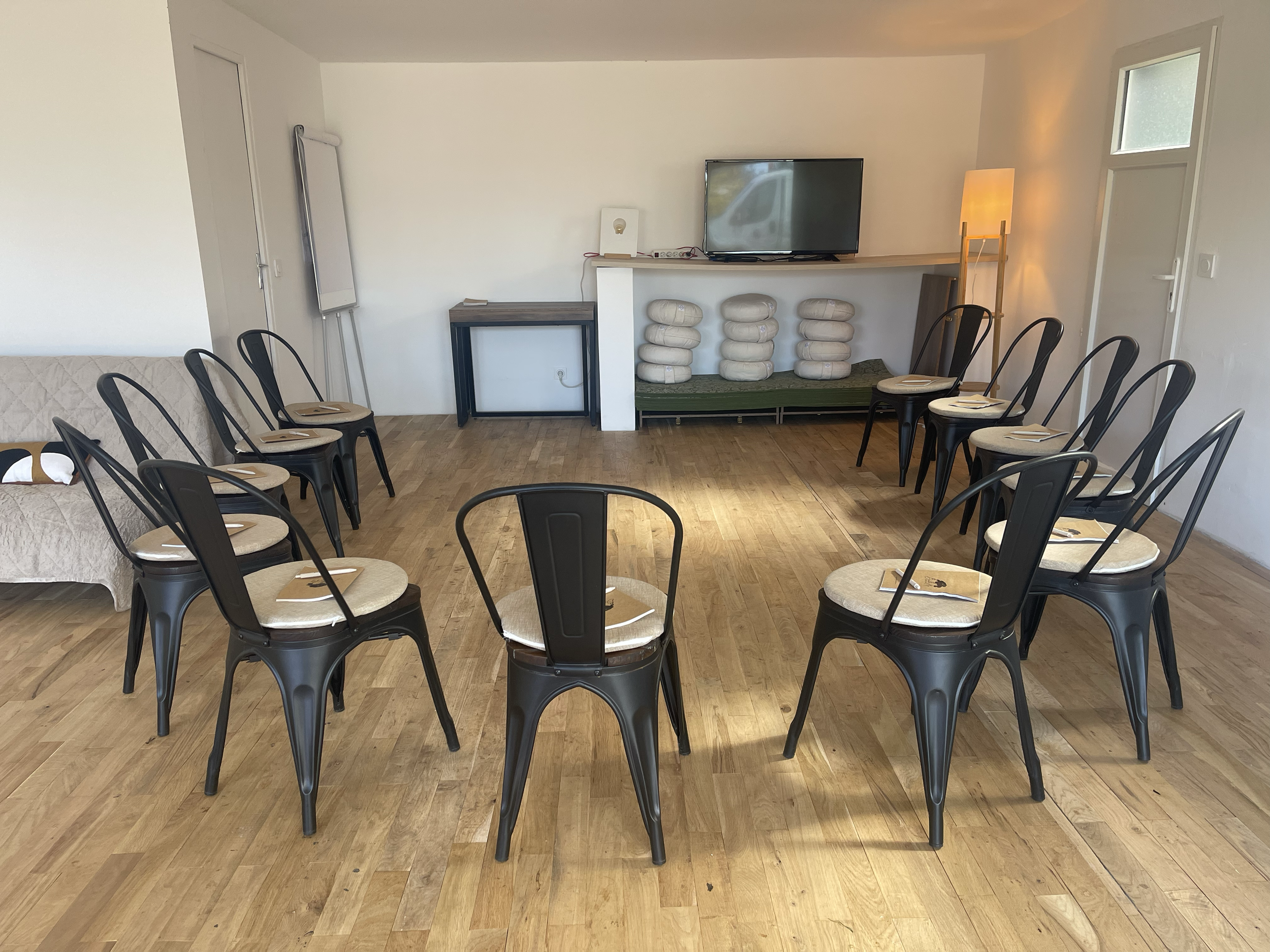 Salle de réunion ou d'événement avec des sièges noirs autour d'une table, un mur blanc avec un petit meuble et une télévision montée, un lampadaire et des fenêtres.