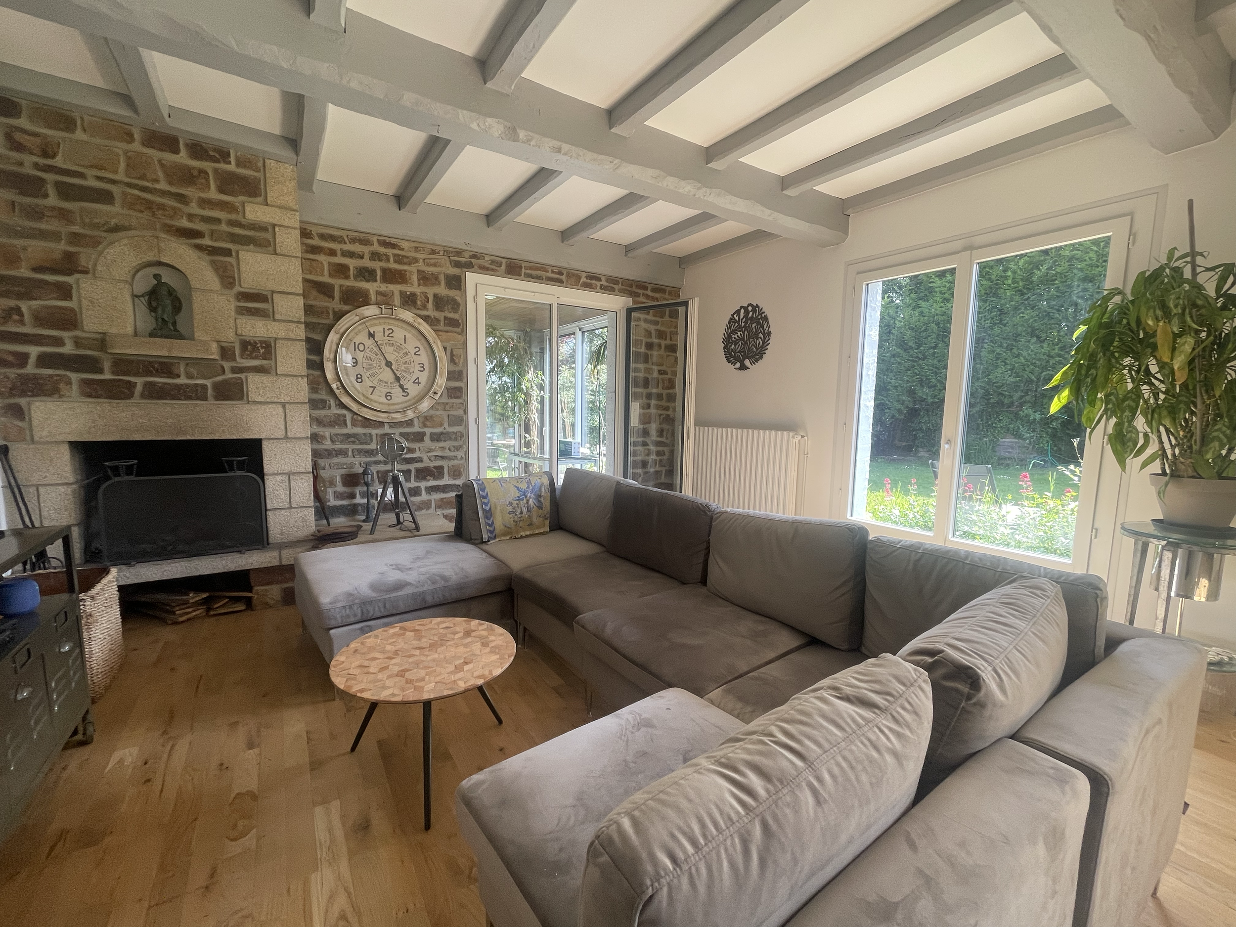 Salon avec un canapé gris, une table basse en bois, un mur en briques avec une horloge et une niche avec une statue, une cheminée en pierre, une grande fenêtre laissant entrer la lumière naturelle, et une plante verte dans un pot blanc.