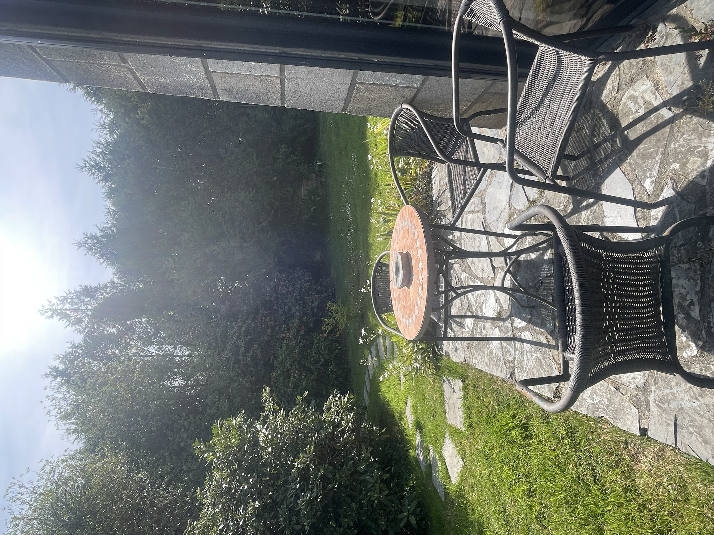 Balcon avec deux chaises en métal noir et une table en mosaïque, terrasse pavée en pierre, jardin avec pelouse et arbustes, ciel clair.