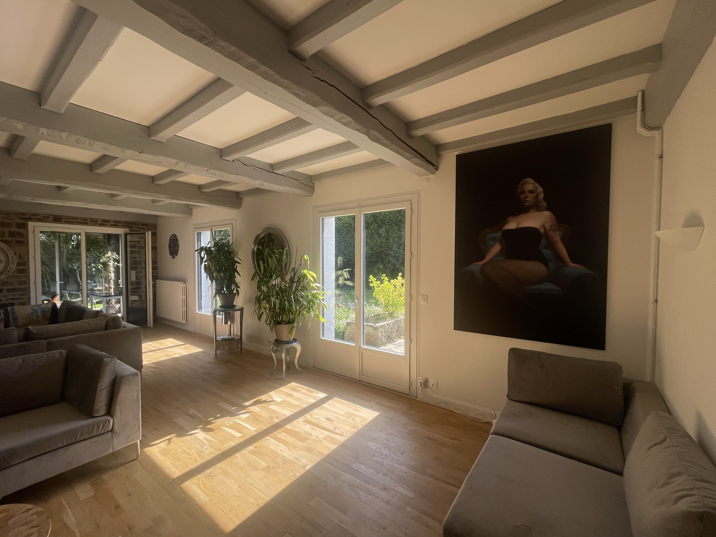 Salon avec plusieurs canapés, grande fenêtre laissant entrer la lumière, tableau de femme sur le mur, et plusieurs plantes d'intérieur.
