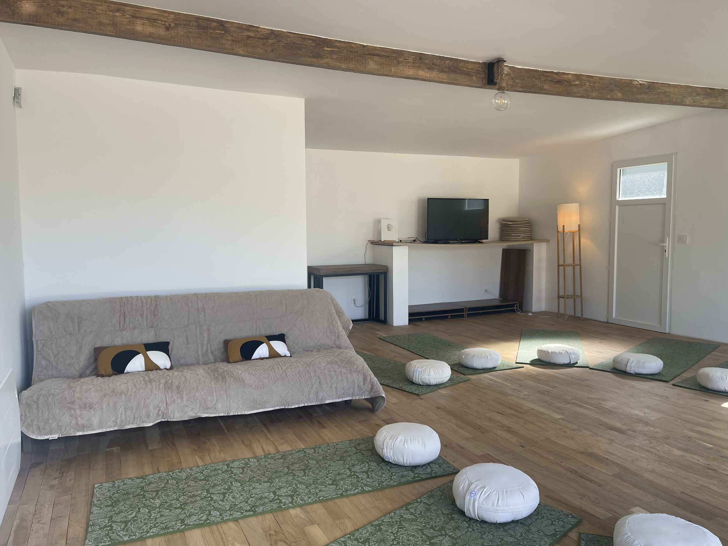 Salon avec un sofa beige recouvert d'une housse, coussins avec motifs abstraits, un meuble en bois sombre, une télévision, une lampe sur une table, des tapis verts avec motifs, des coussins blancs au sol, une porte fenêtre et un plafond avec poutres 