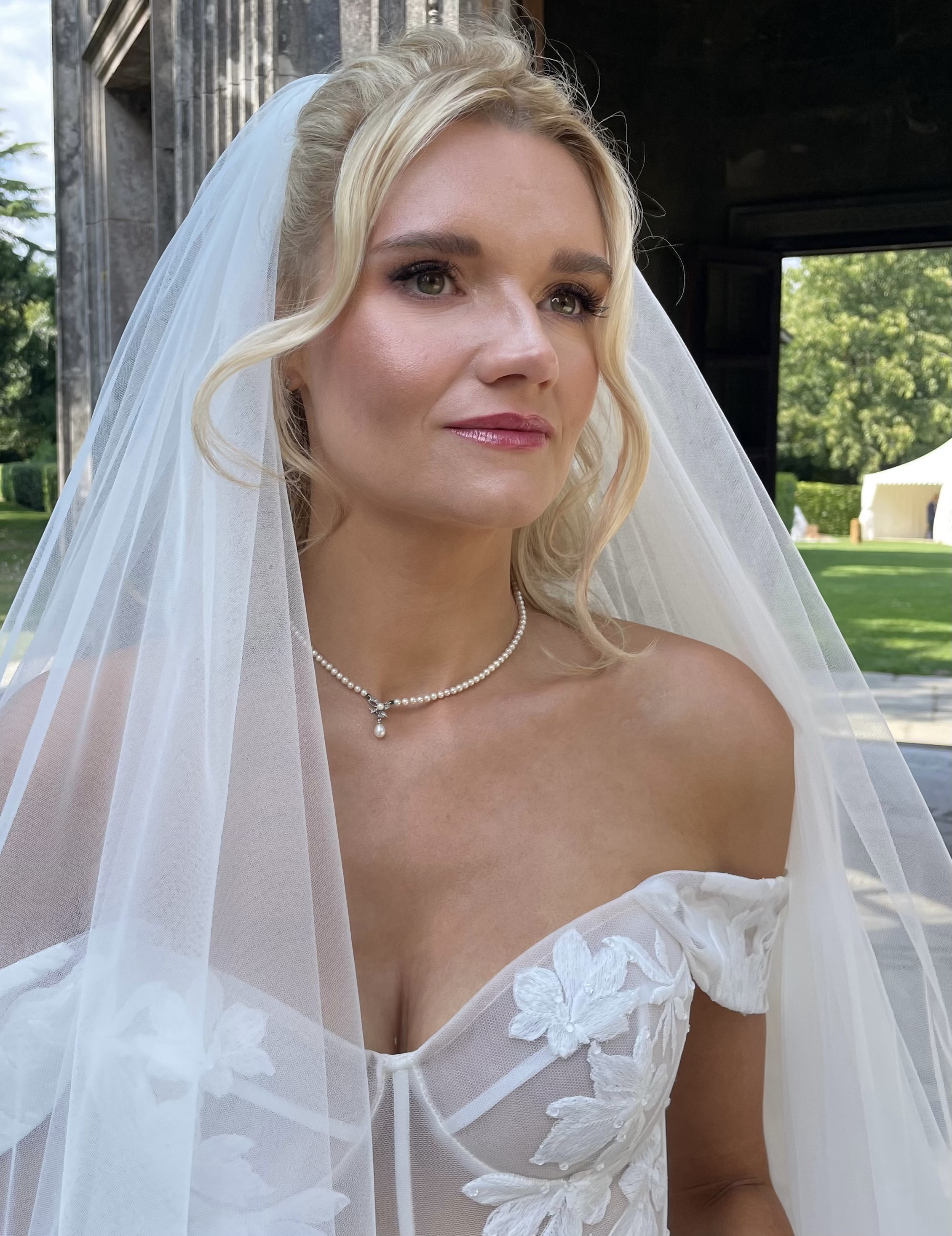 Close-up of a bride with blonde, softly waved hair,  wearing a white wedding dress with floral embroidery, a pearl necklace, and a sheer veil, outdoors with green trees and a tent in the background. beautifully glowing skin, plum eyeliner