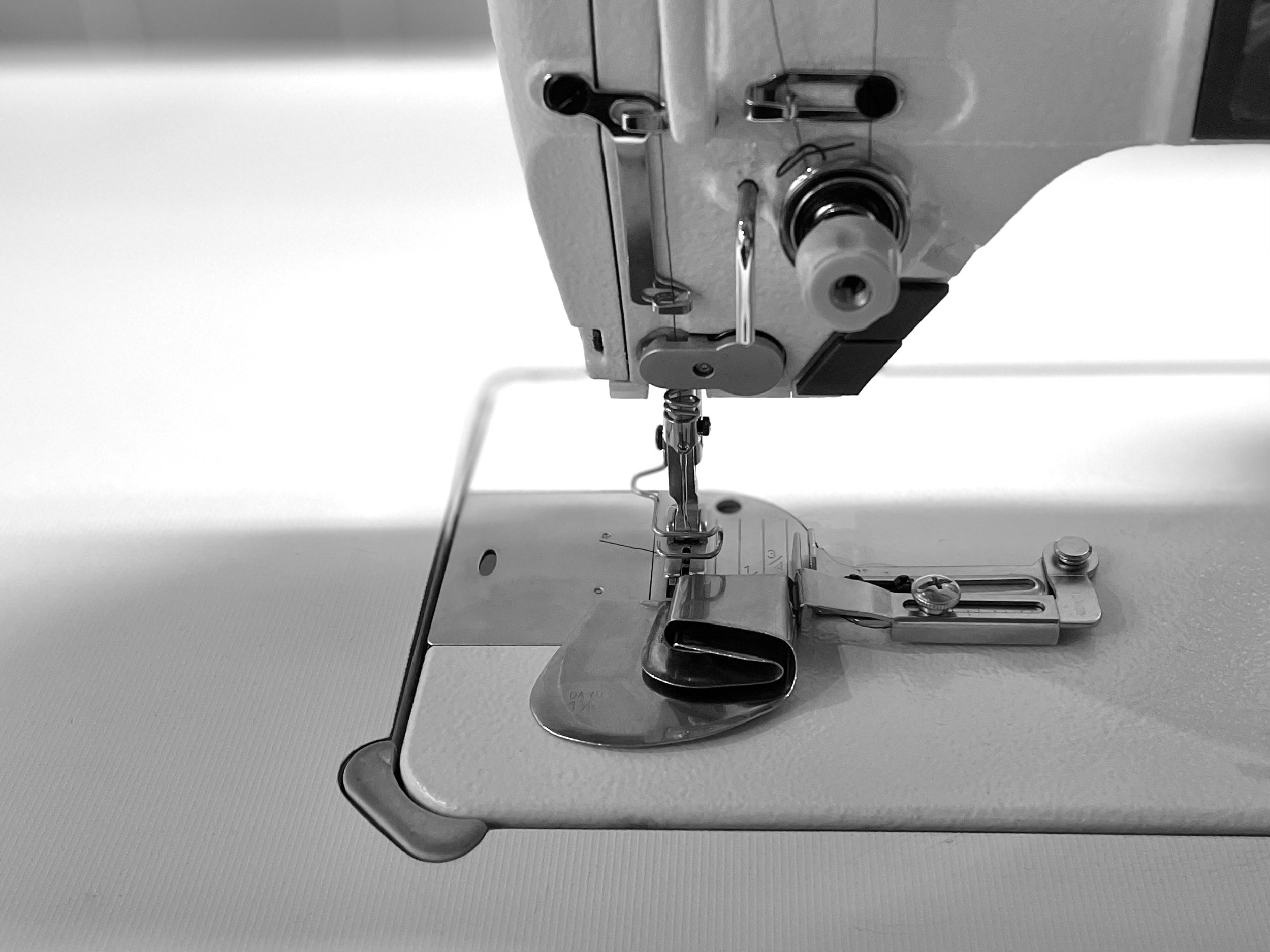 Close-up of a sewing machine's needle, presser foot, and feed dogs against a plain background. DAW