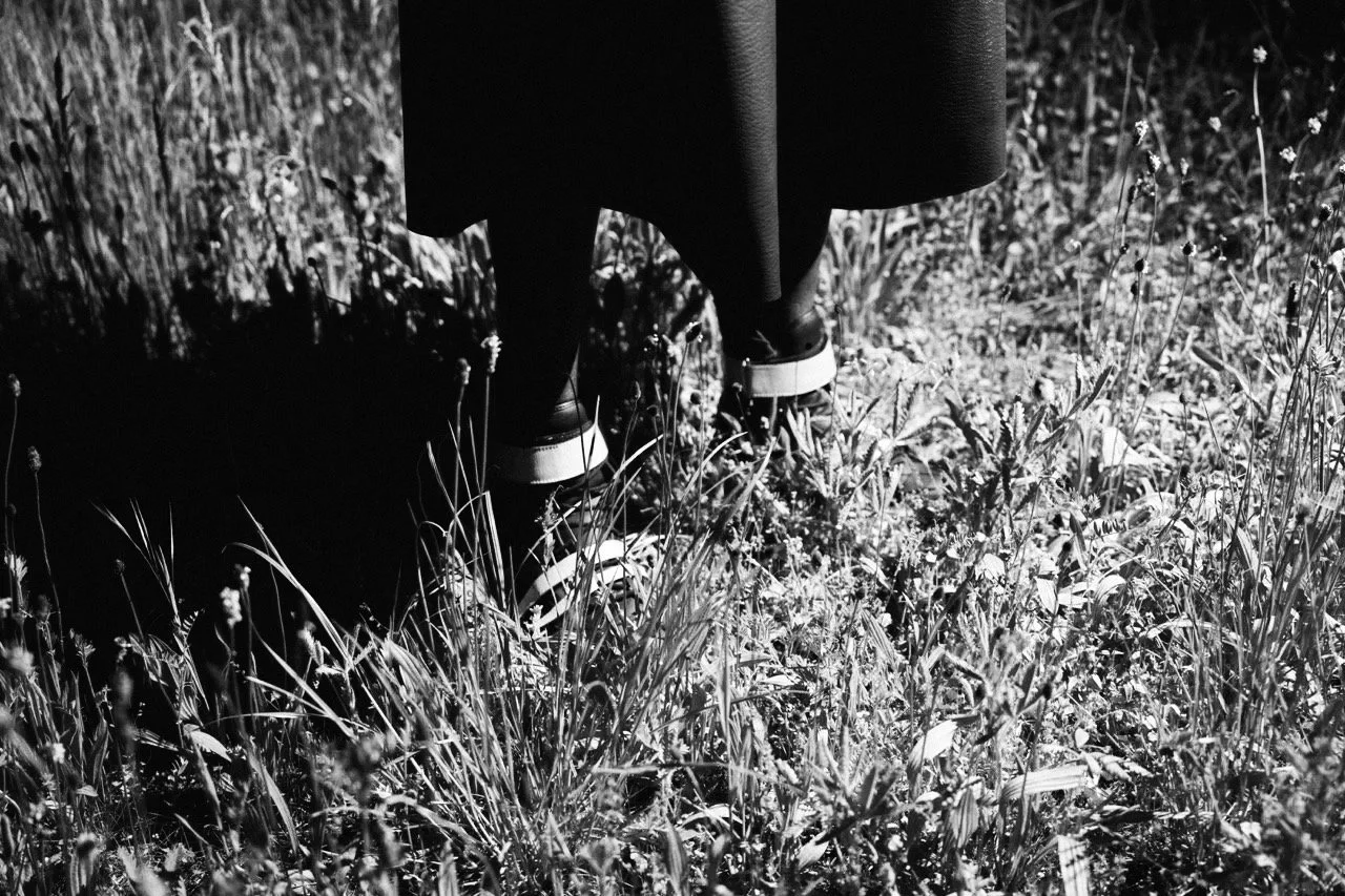 Lower half of a person in oversized leather shorts and black sneakers standing in a grassy field with wildflowers, black and white photo. Daniel Alexander Weiter - DAW