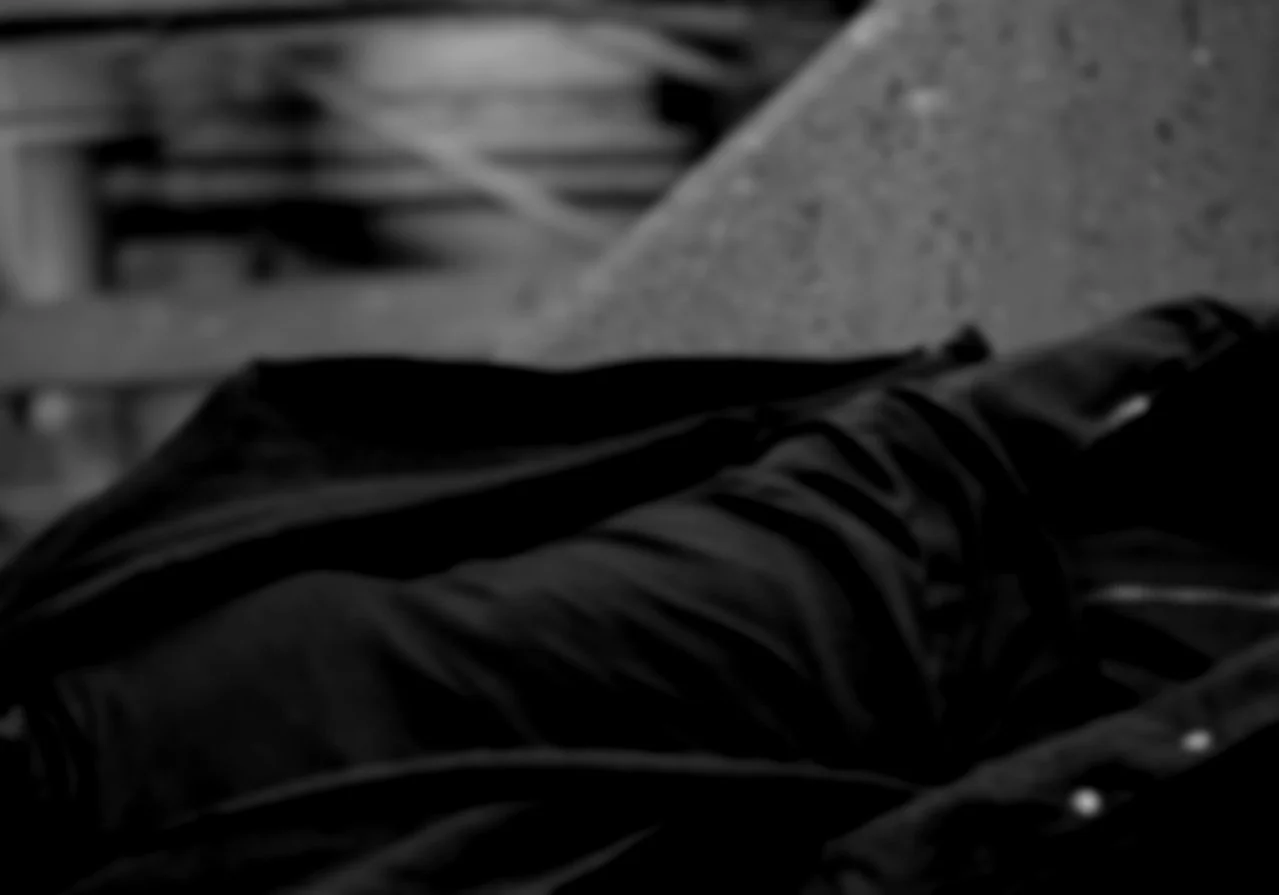 A black and white photo showing a person's lap with dark pants, with a blurred background of wooden structures or pallets. Daniel Alexander Weiter - DAW