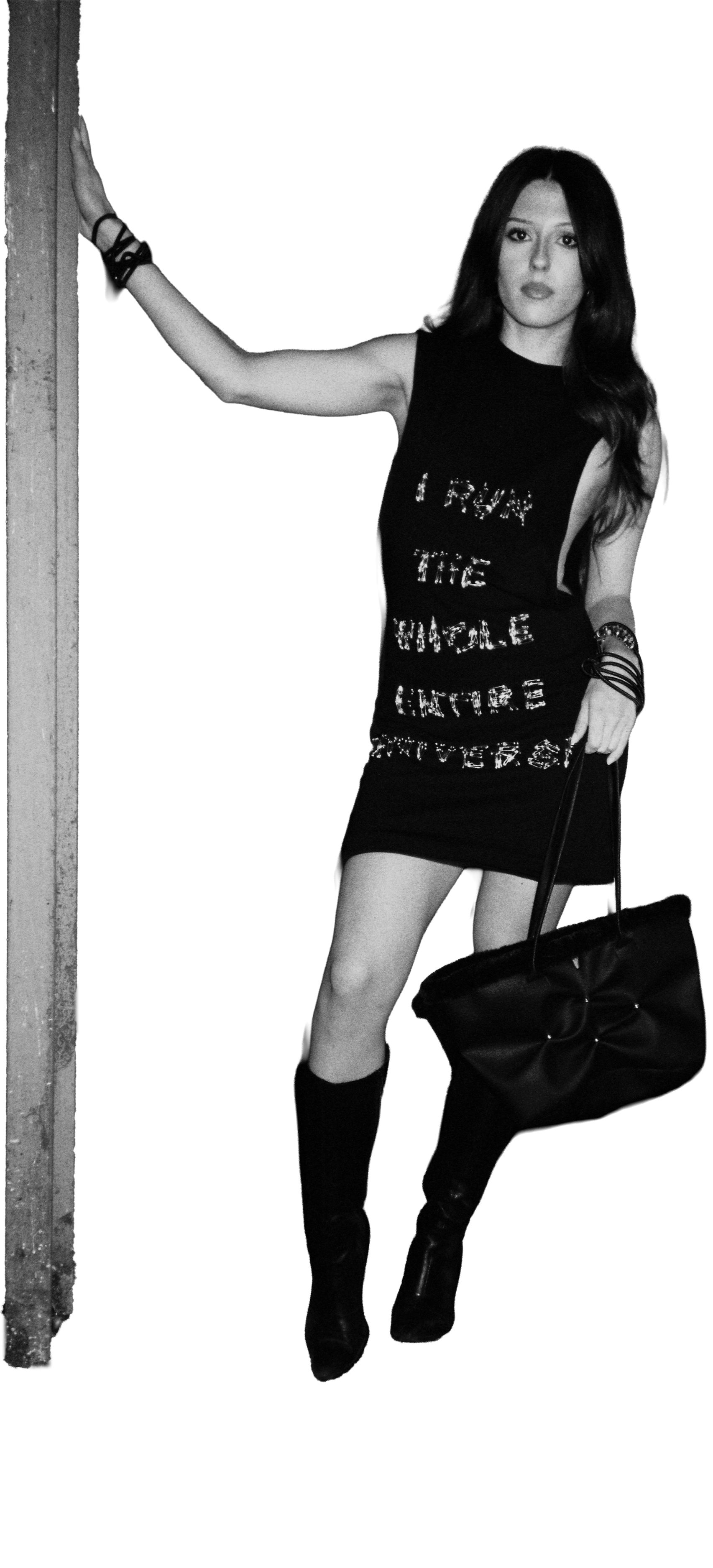 Black and white photo of a woman standing against a dark background, wearing a sleeveless black  Miss Universe dress with embroidered text out of safety pins, black high boots, and holding a large black Ripple bag. Daniel Alexander Weiter - DAW