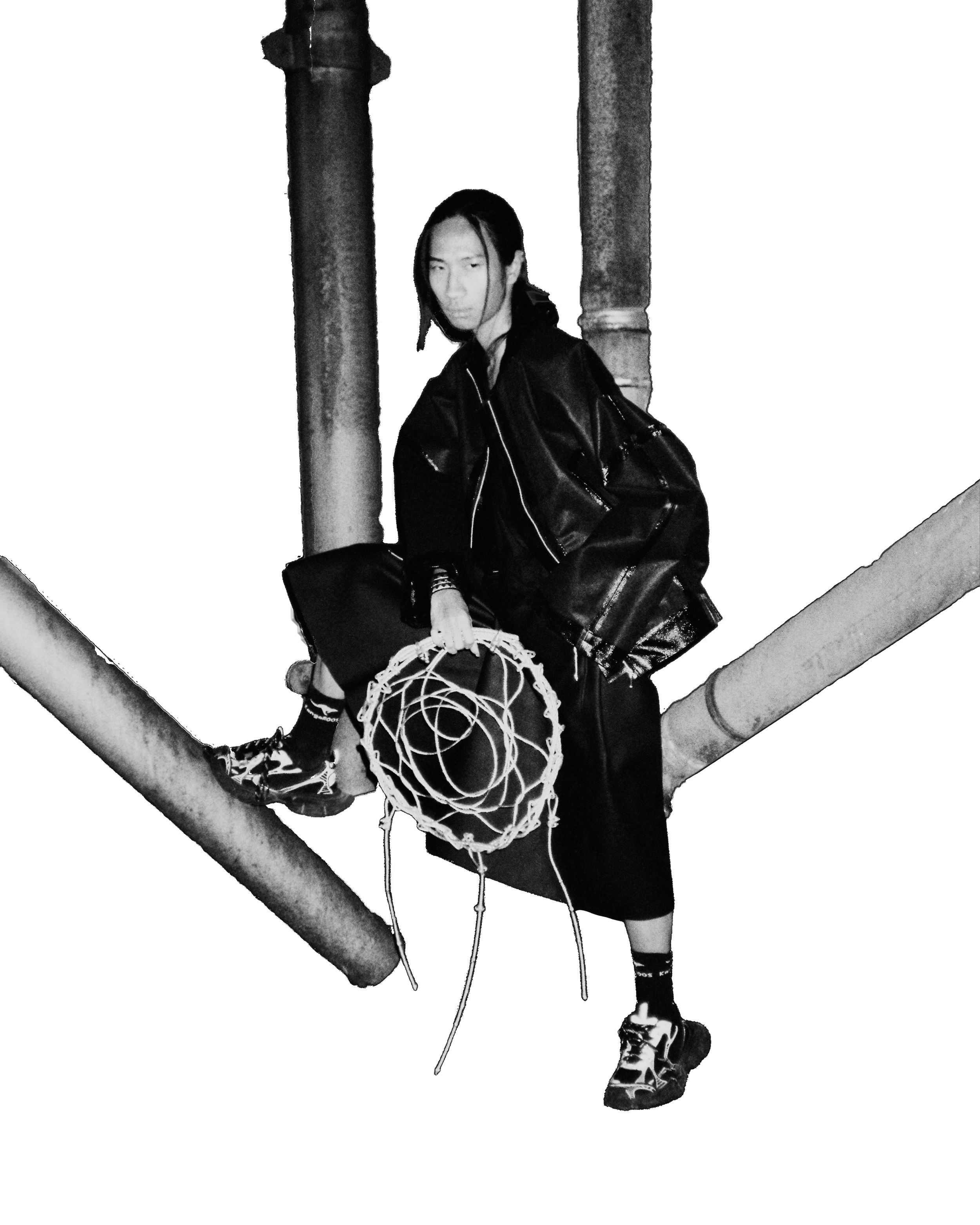 A person with long hair wearing a dark jacket and patterned shoes, standing on a structure with pipes, holding a large metal Dreamcatcher, in a dark brutalistic setting. Daniel Alexander Weiter - DAW